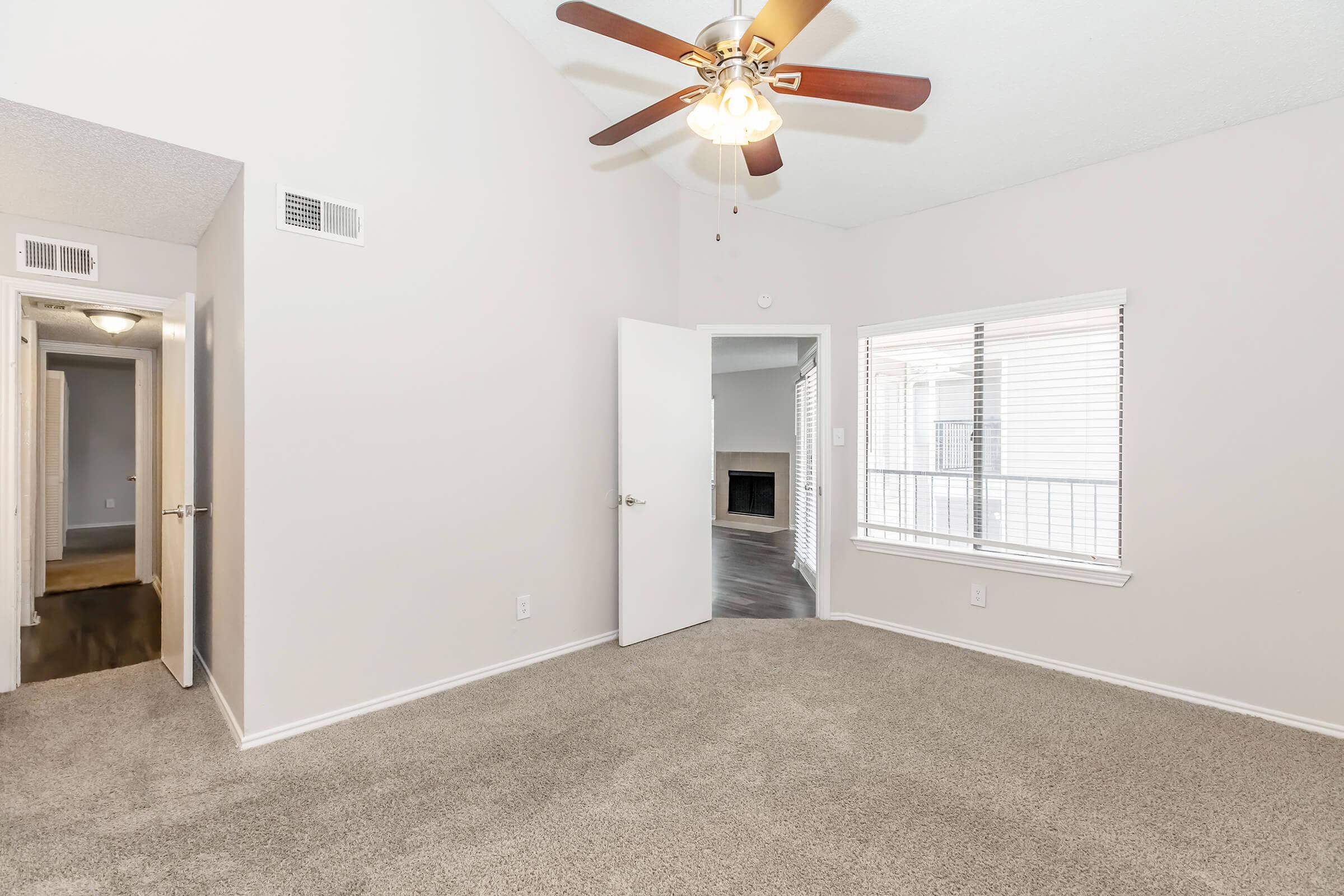 A spacious, well-lit room with beige carpet, ceiling fan, and neutral-colored walls. There is a doorway leading to a hallway on the left and a large window with a view outside. The right side features a fireplace, adding a cozy touch to the space.