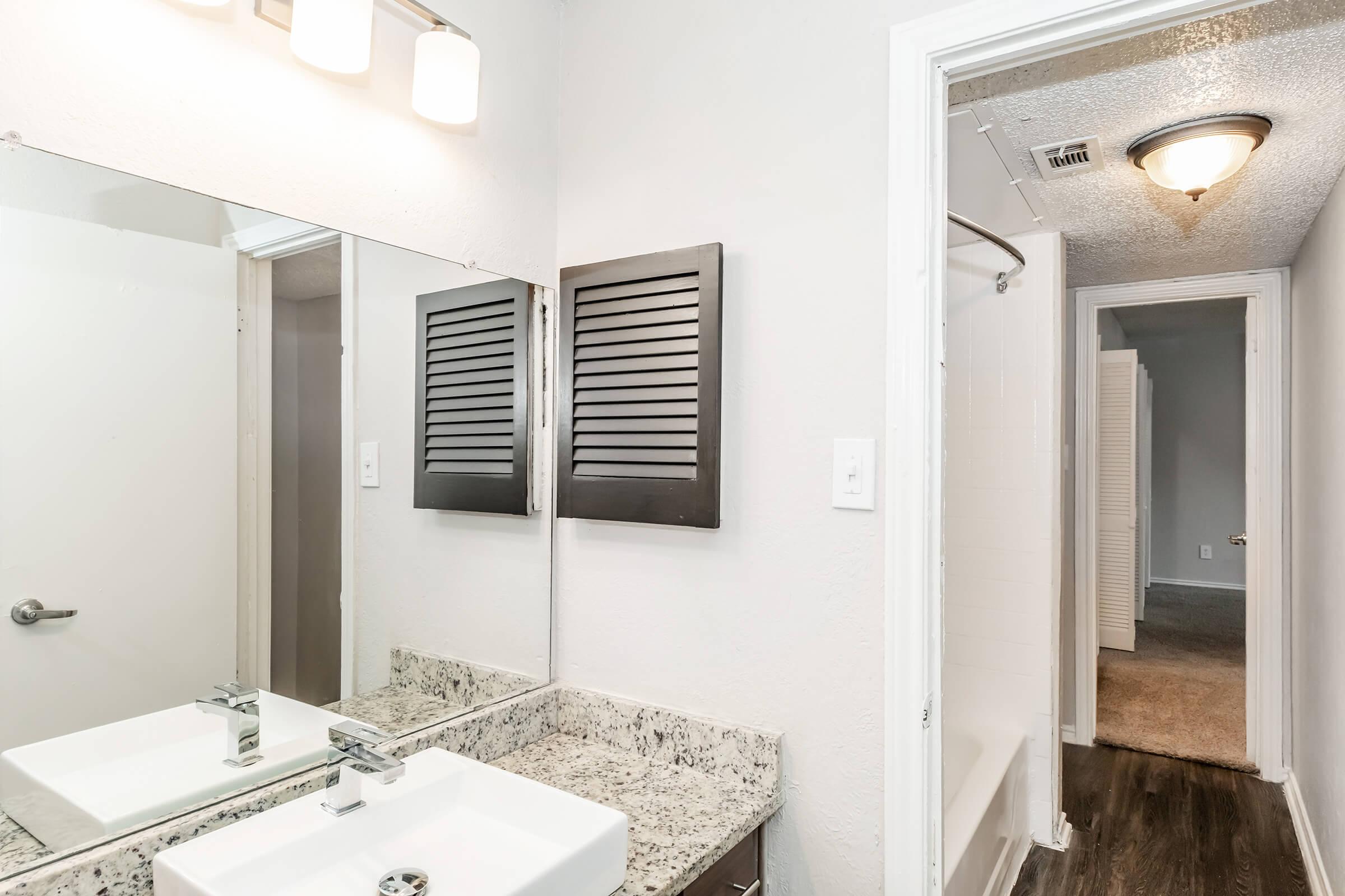 A modern bathroom with a granite countertop and a rectangular sink. Above the sink, there are three light fixtures. The wall mirror is framed, and to the side are decorative louvered panels. A doorway in the background leads to a carpeted hallway. The overall color scheme is neutral and contemporary.