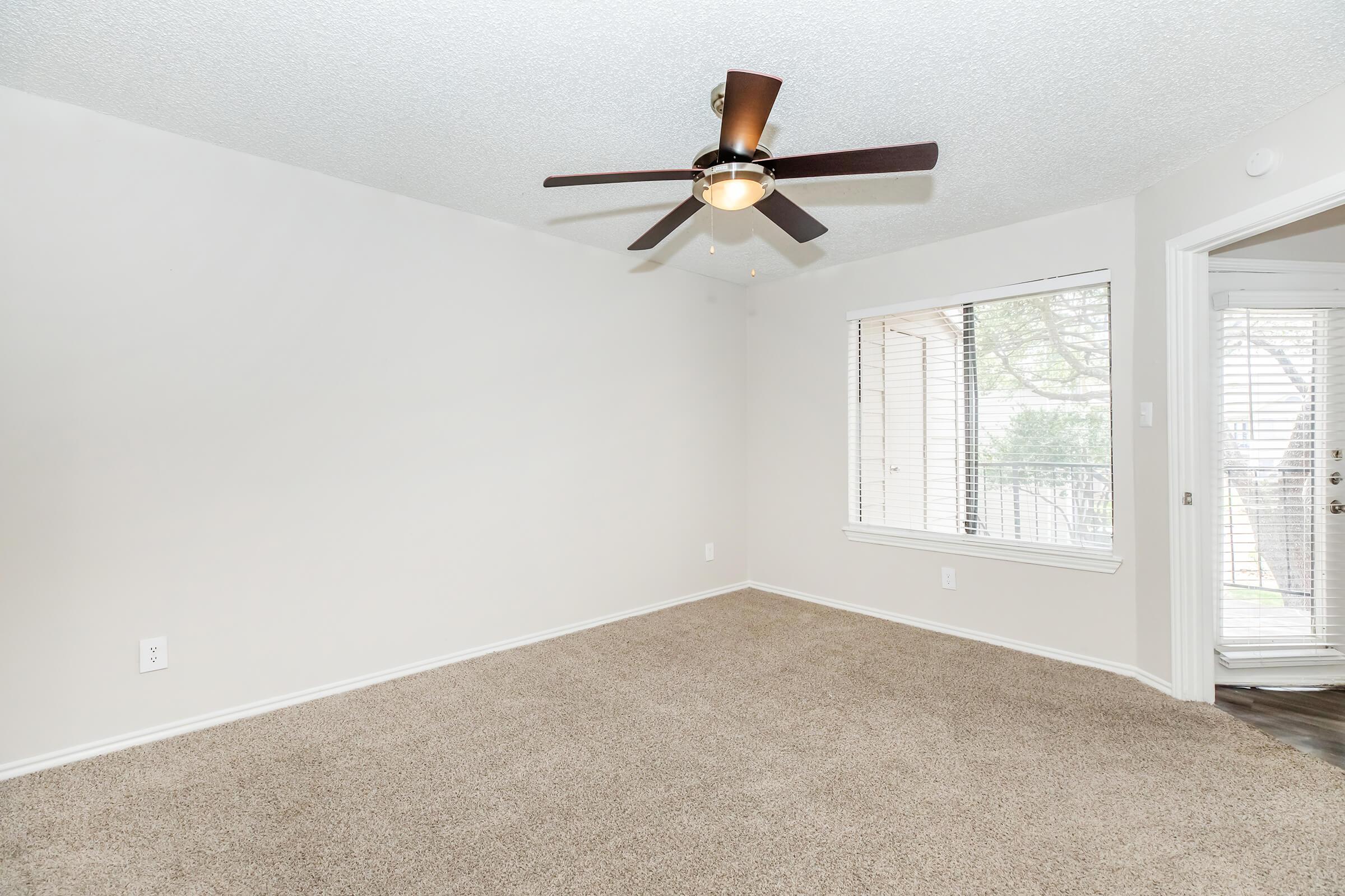 Empty room featuring beige carpet flooring, a ceiling fan with wooden blades, and a large window with blinds. The walls are painted a light color, and there is a door leading outside to a small balcony. Natural light enters the room, creating a bright and airy atmosphere.