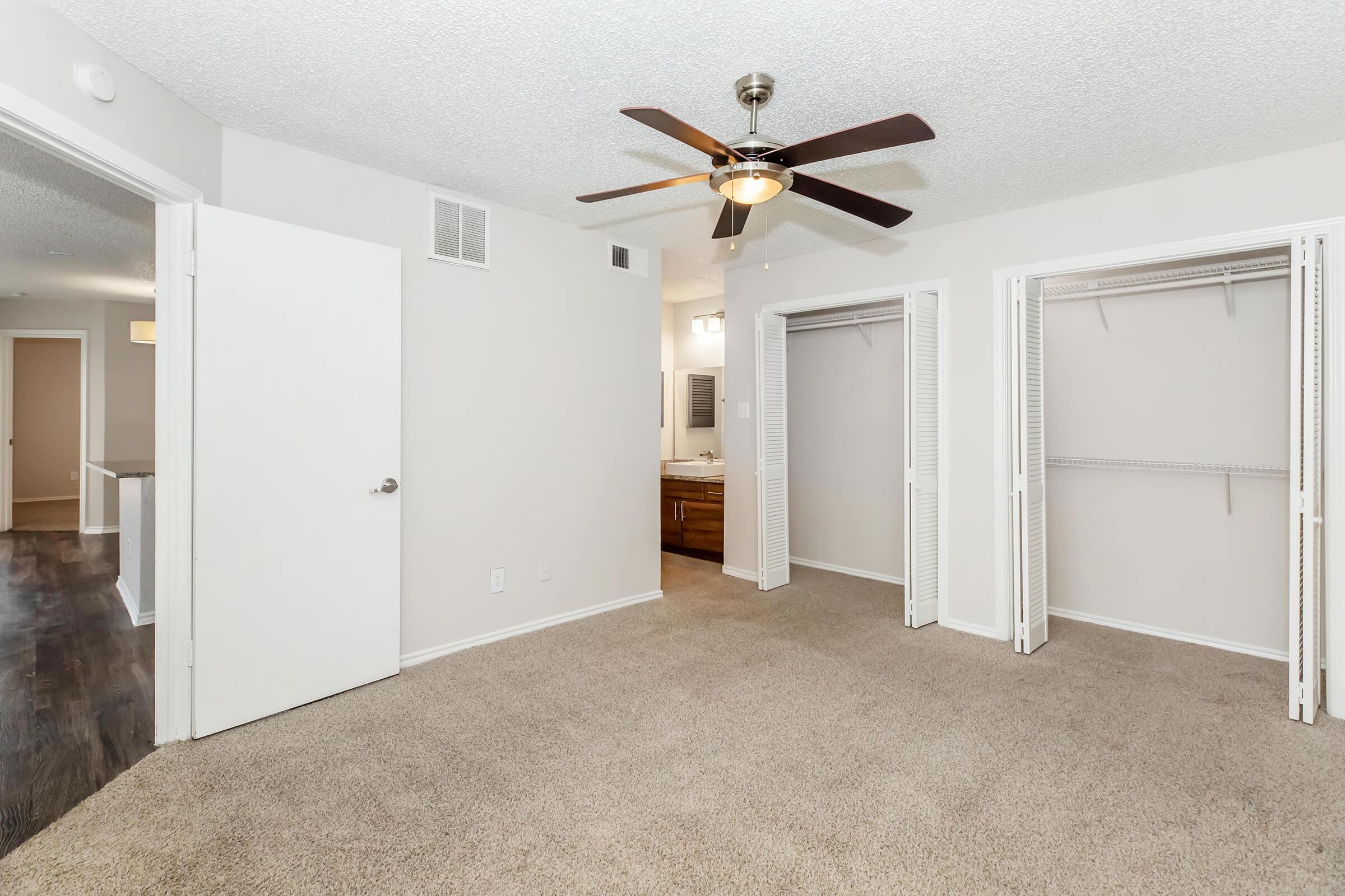 Spacious bedroom featuring beige carpet, a ceiling fan, and dual closets with sliding doors. A doorway leads to a bathroom area, and light-colored walls enhance the room's brightness. The overall layout shows an open design with natural light flowing in.