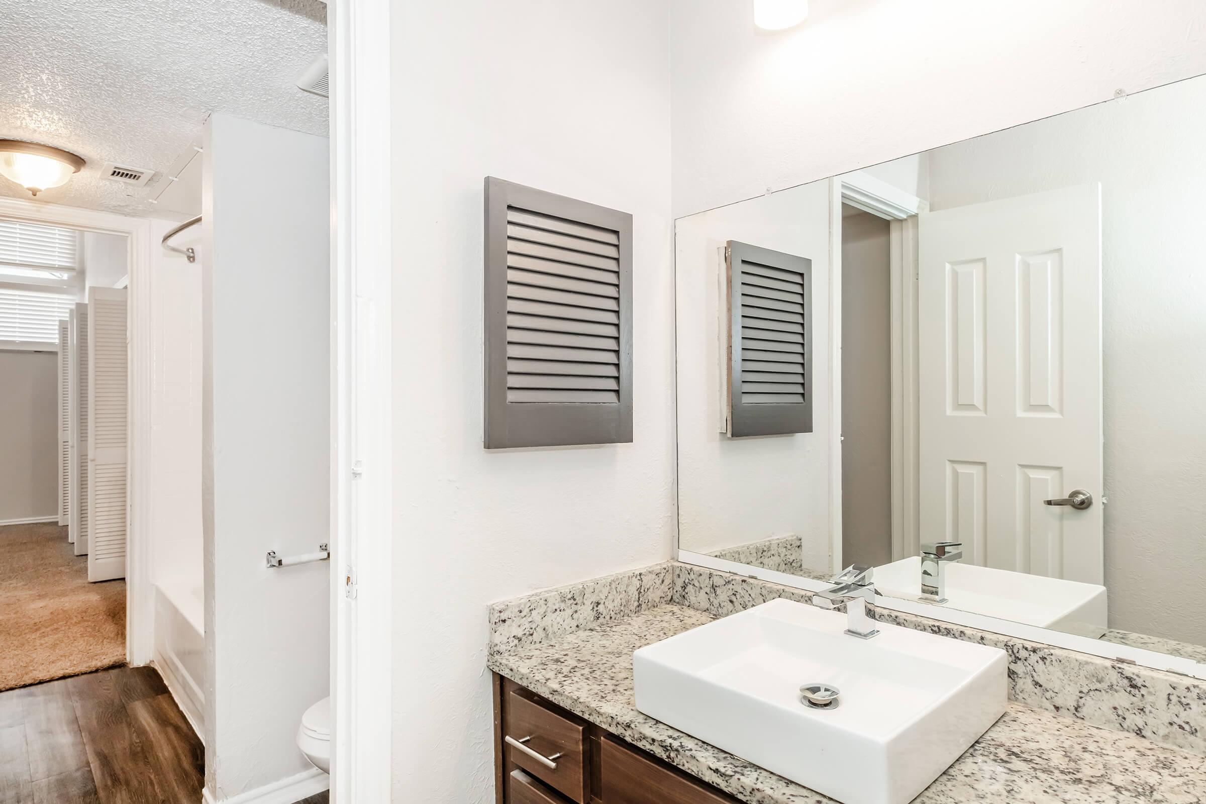 A modern bathroom featuring a sleek vanity with a square sink, granite countertop, and a large mirror. A door leads to a separate area, and there is a shower stall visible in the background. The walls are painted in a light color, enhancing the bright and spacious feel of the room.