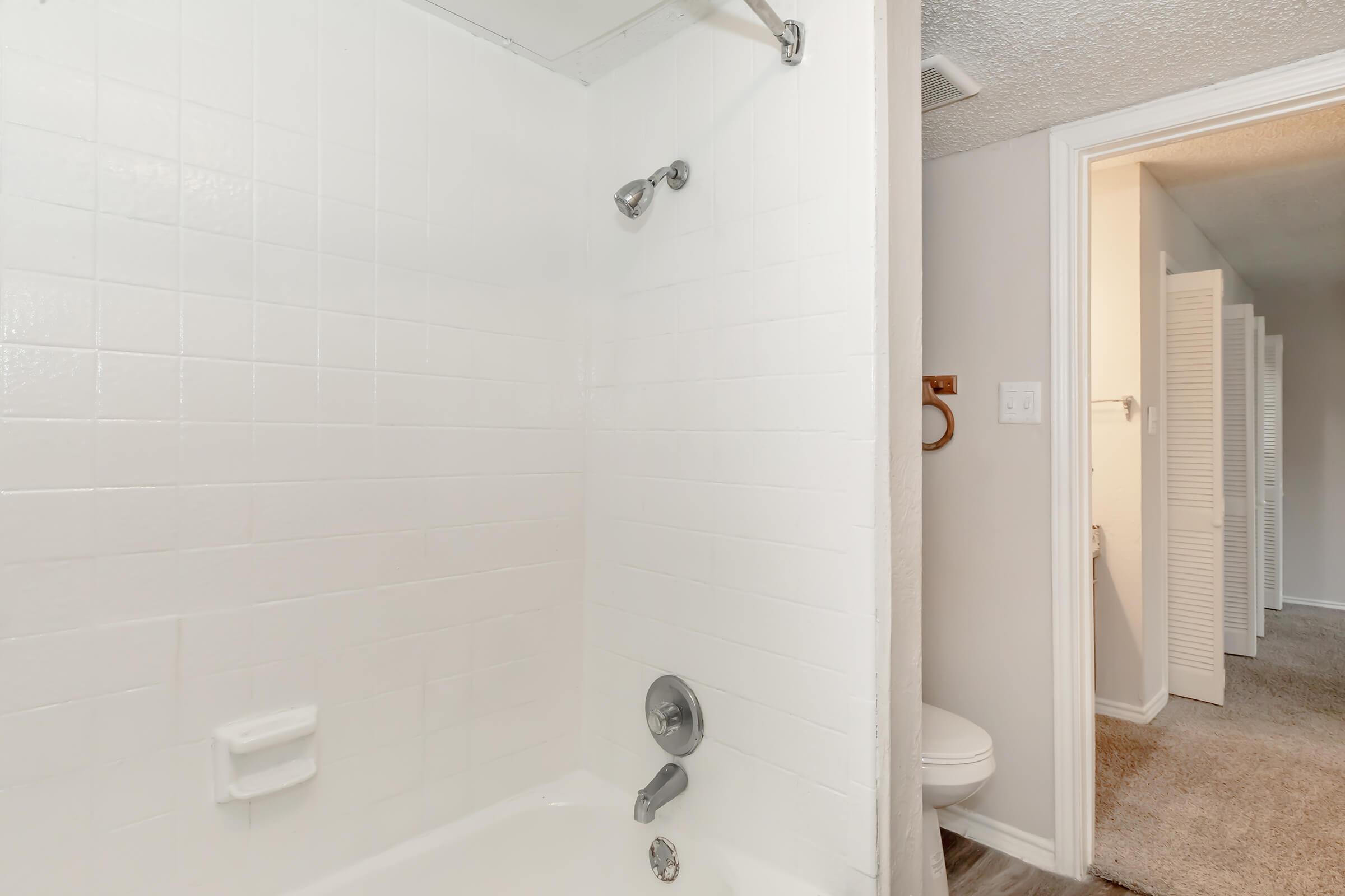 A clean and simple bathroom featuring a white tiled shower with a sliding glass door. The shower has a modern chrome showerhead and faucet. In the background, a glimpse of a hallway and a bathroom door can be seen, along with a light-colored carpeted floor.