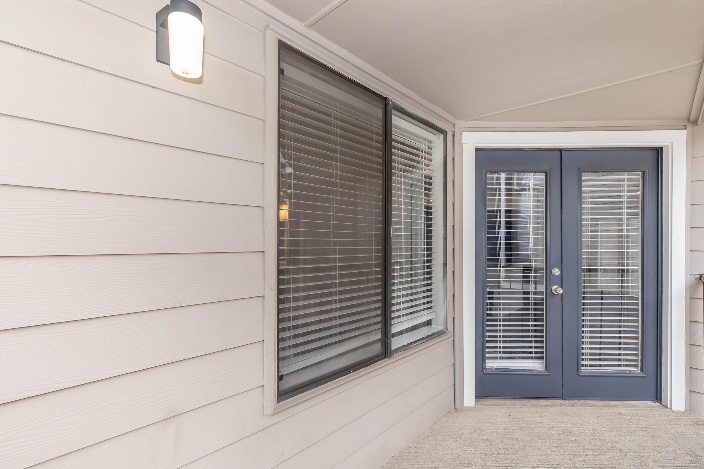 A front entrance with a double door on the right, featuring vertical blinds on the adjacent window. The wall is light-colored wood, and there is an outdoor wall light fixture above the doorway. The area has a clean, modern aesthetic.