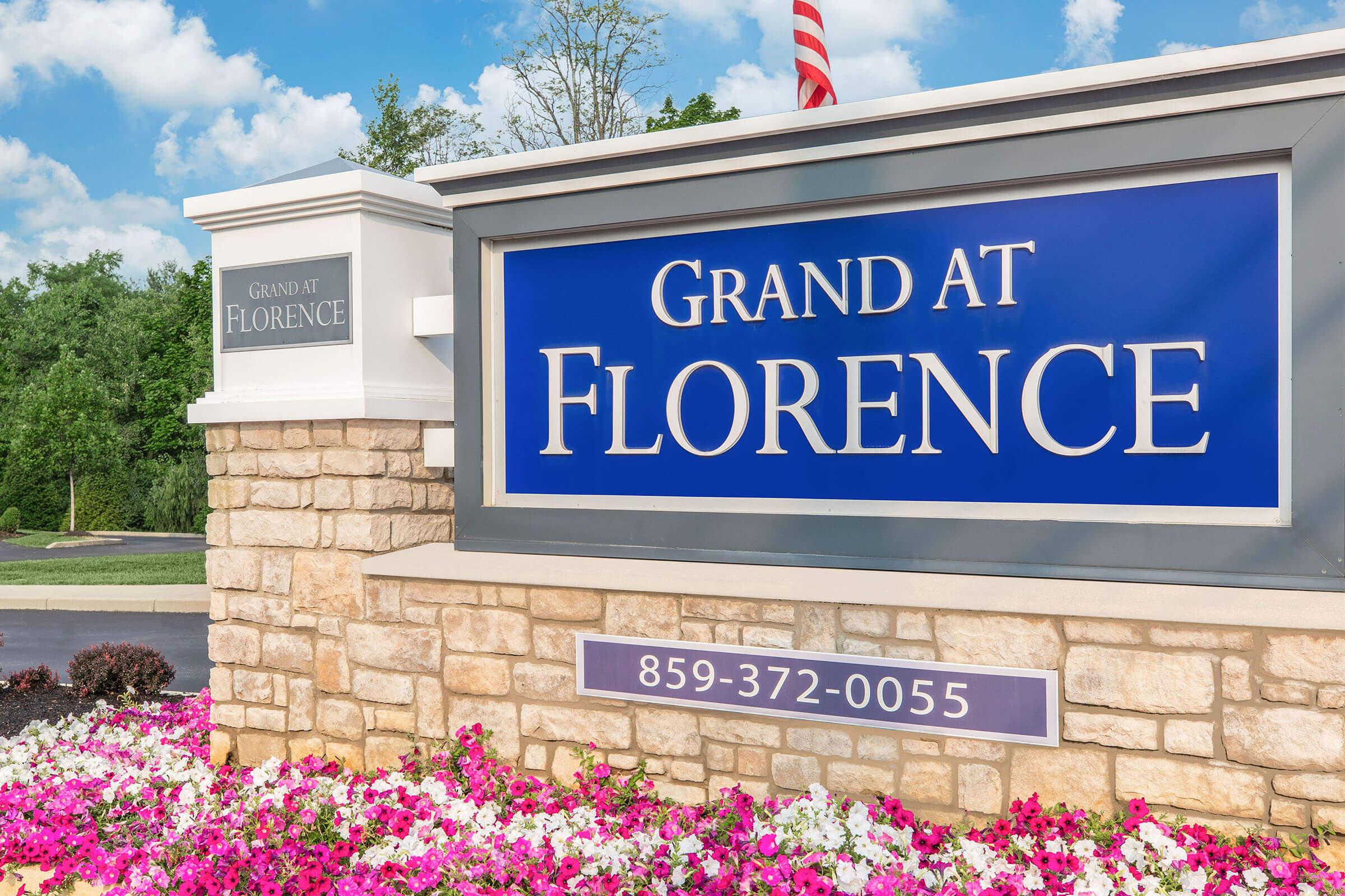 Sign reading "Grand at Florence" with a stone base and colorful flowers in front, under a blue sky.