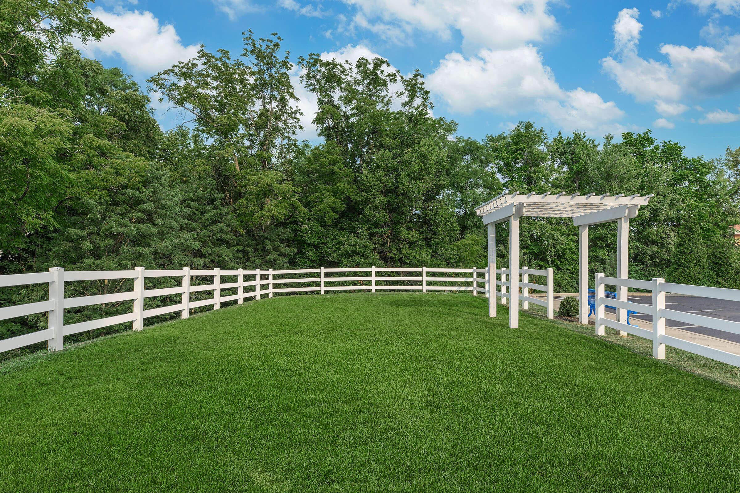 A landscaped area featuring a lush green lawn bordered by a white wooden fence, with a pergola structure at one end and trees in the background under a partly cloudy sky.