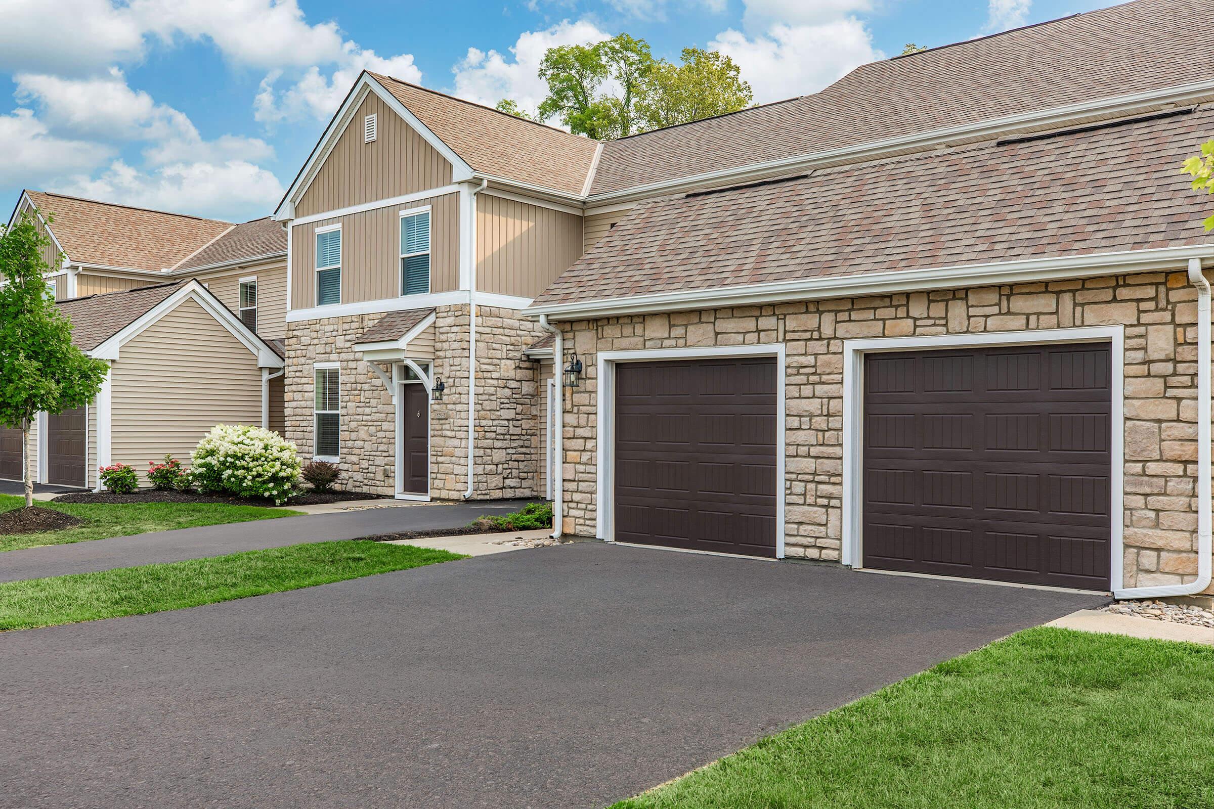 A modern residential building with a combination of beige siding and stone accents. The front features a symmetrical design with windows and a double garage door. A well-maintained grassy area and a few shrubs are visible in the front yard, with a clear blue sky and fluffy clouds in the background.