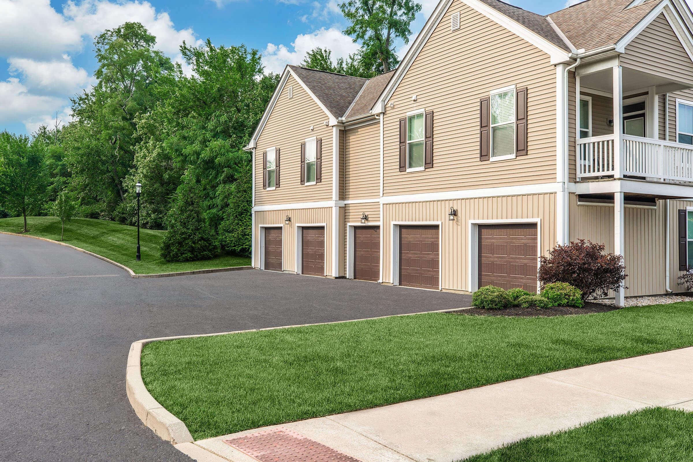 A residential area featuring a two-story building with attached garages, surrounded by well-maintained lawns and trees, under a partly cloudy sky. The pavement is clean and the landscape is green and inviting.