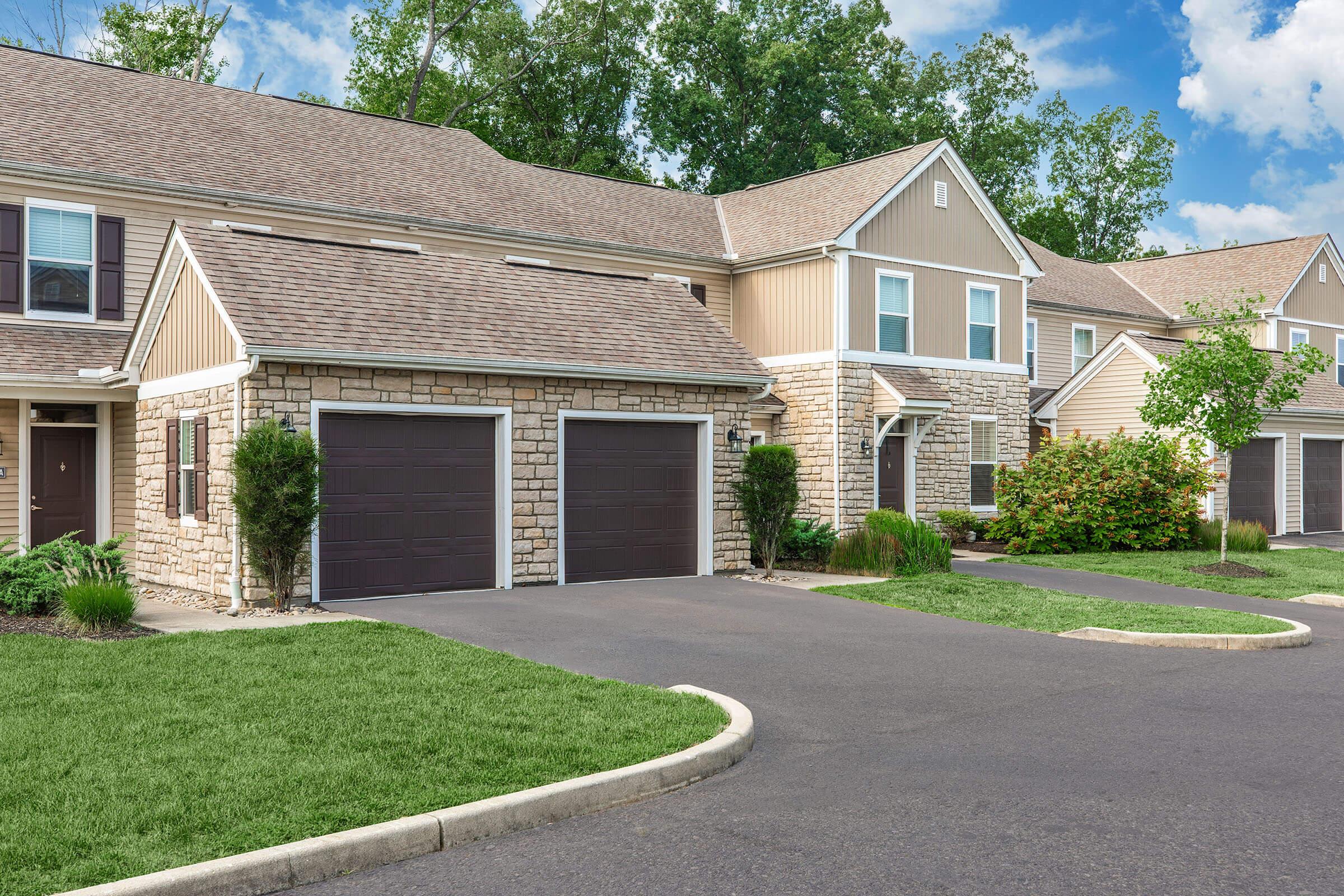 Residential building featuring a two-car garage, a stone and siding exterior, and well-maintained landscaping with green grass and bushes. The sky is clear with scattered clouds.