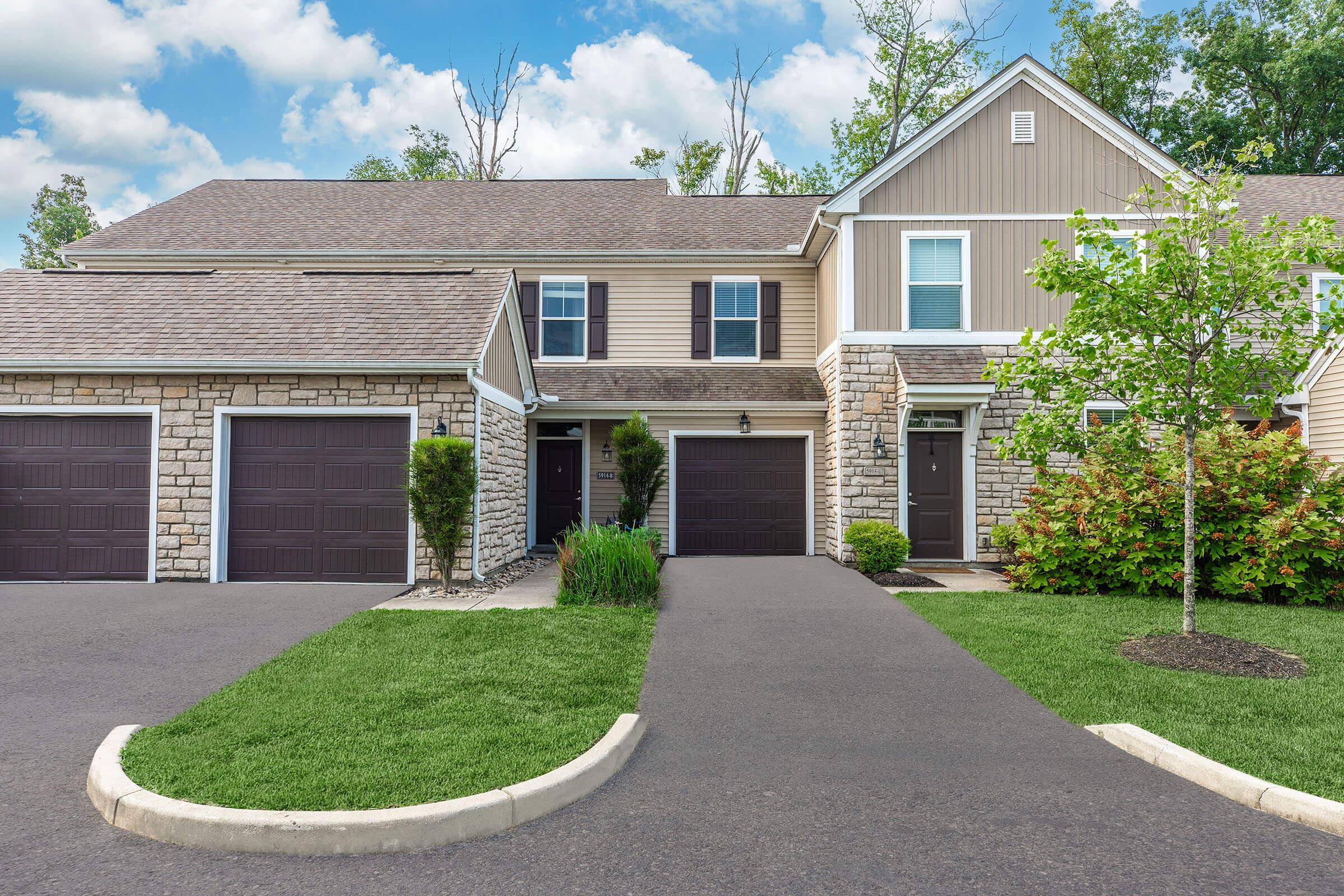 A modern two-story townhouse with a stone and siding exterior, featuring a driveway and well-maintained landscaping. The house on the left has a single garage door, while the one on the right has a slightly different design. Lush green trees and shrubs are visible in the surroundings.