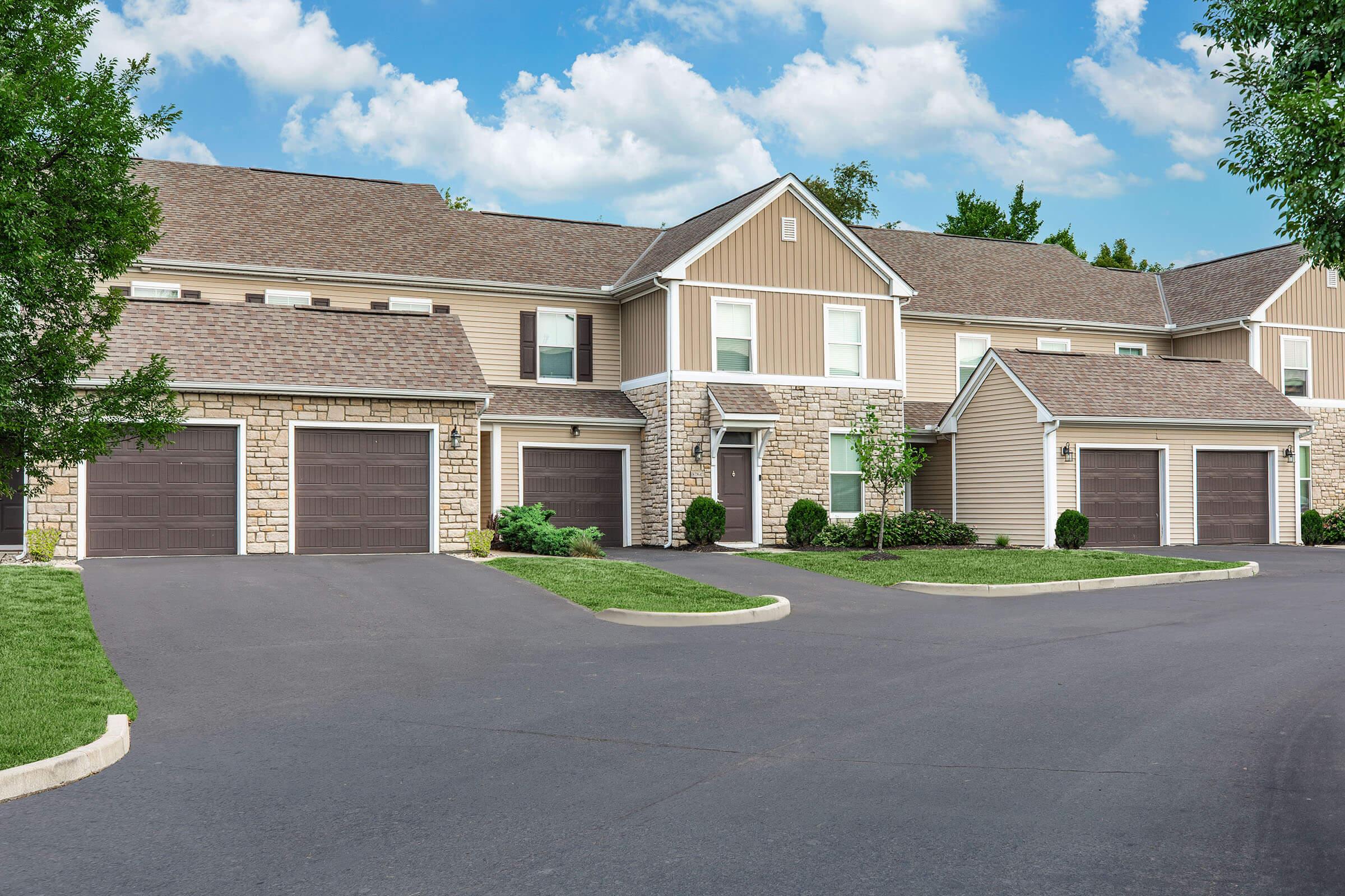 A well-maintained residential building featuring multiple units, with stone and wood siding, a paved driveway, and neatly landscaped lawns under a partly cloudy sky.