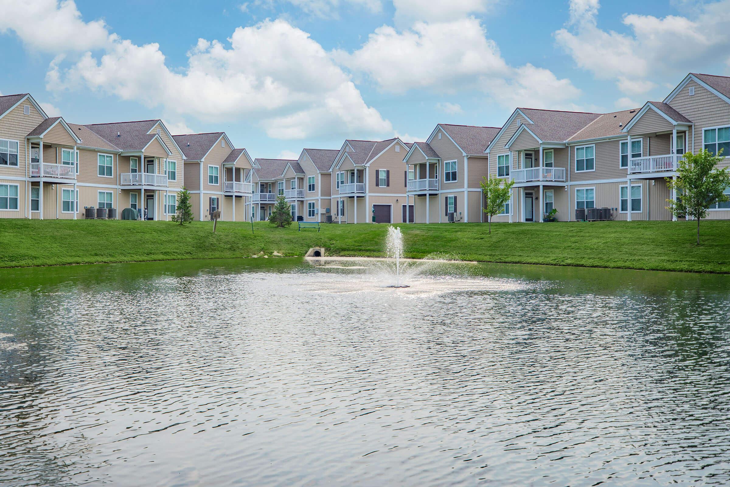 A serene residential area featuring multiple beige apartment buildings situated next to a calm pond with a fountain. The sky is partly cloudy, and green grass surrounds the water, creating a peaceful atmosphere.