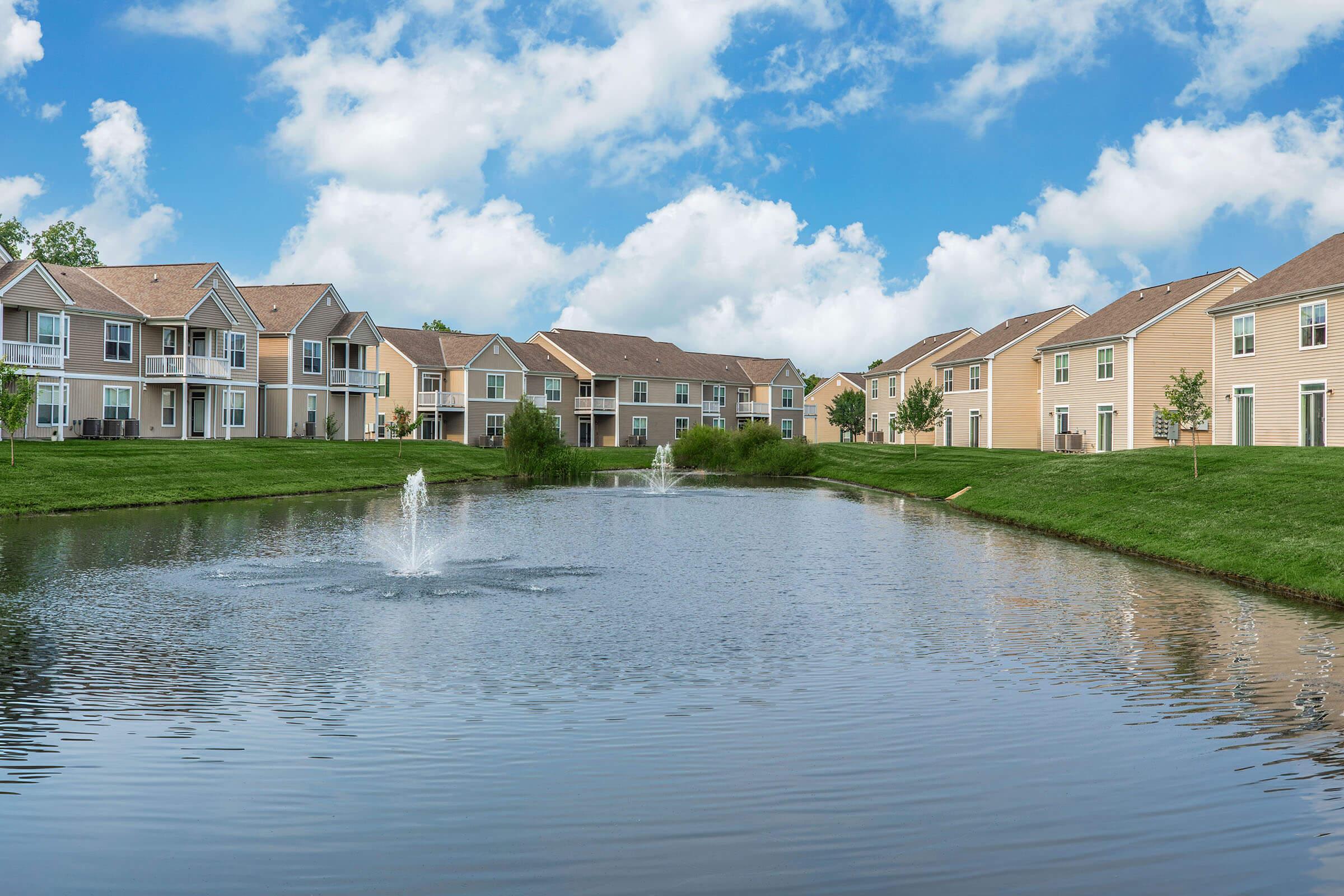 A serene residential area featuring two-story houses with light-colored siding, set along a pond with fountains. The landscape includes green lawns and scattered trees under a bright blue sky with fluffy white clouds.