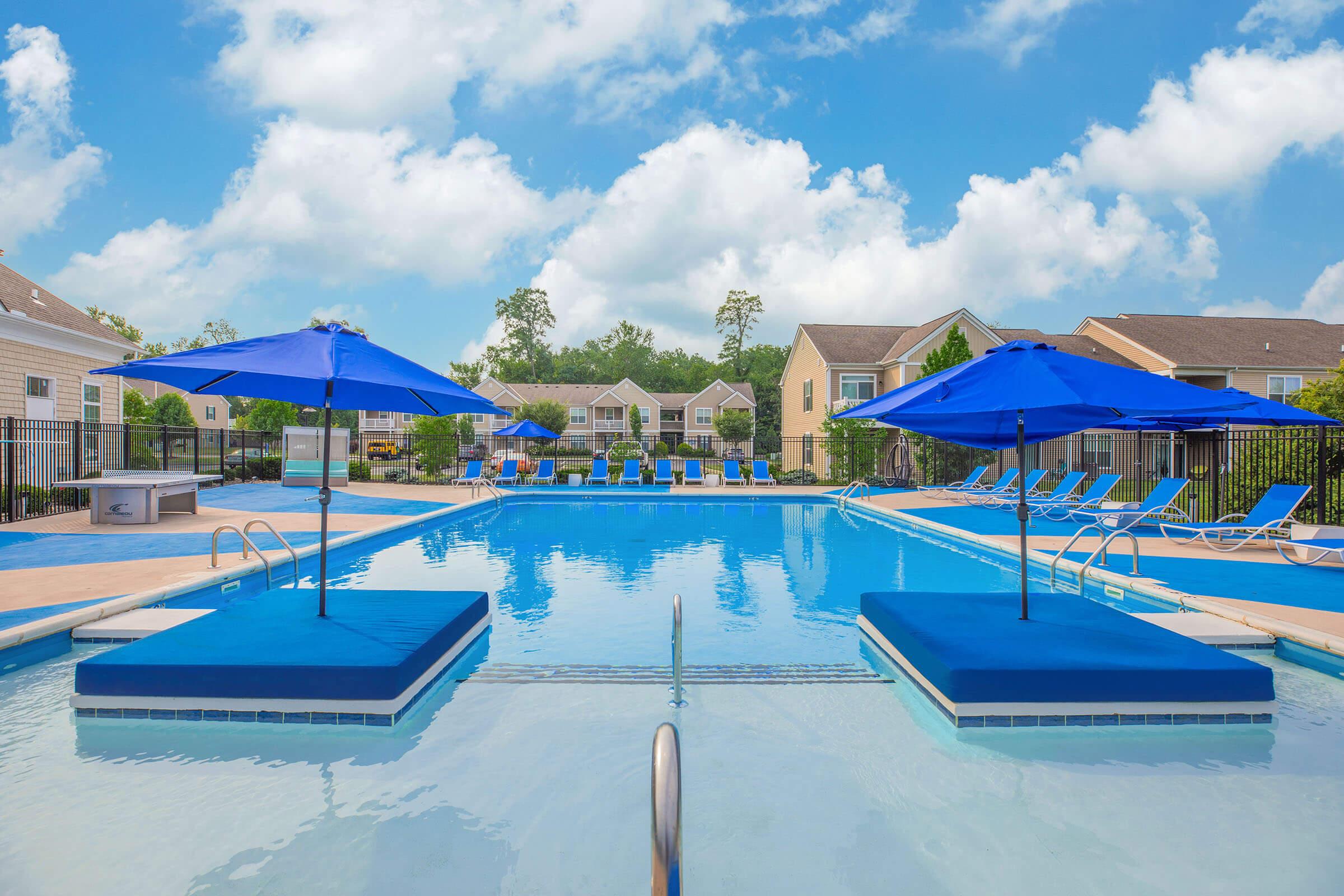 A serene swimming pool scene featuring two large blue umbrellas on floating platforms, surrounded by clear water. In the background, there are lounge chairs and residential buildings, under a bright blue sky with fluffy white clouds.