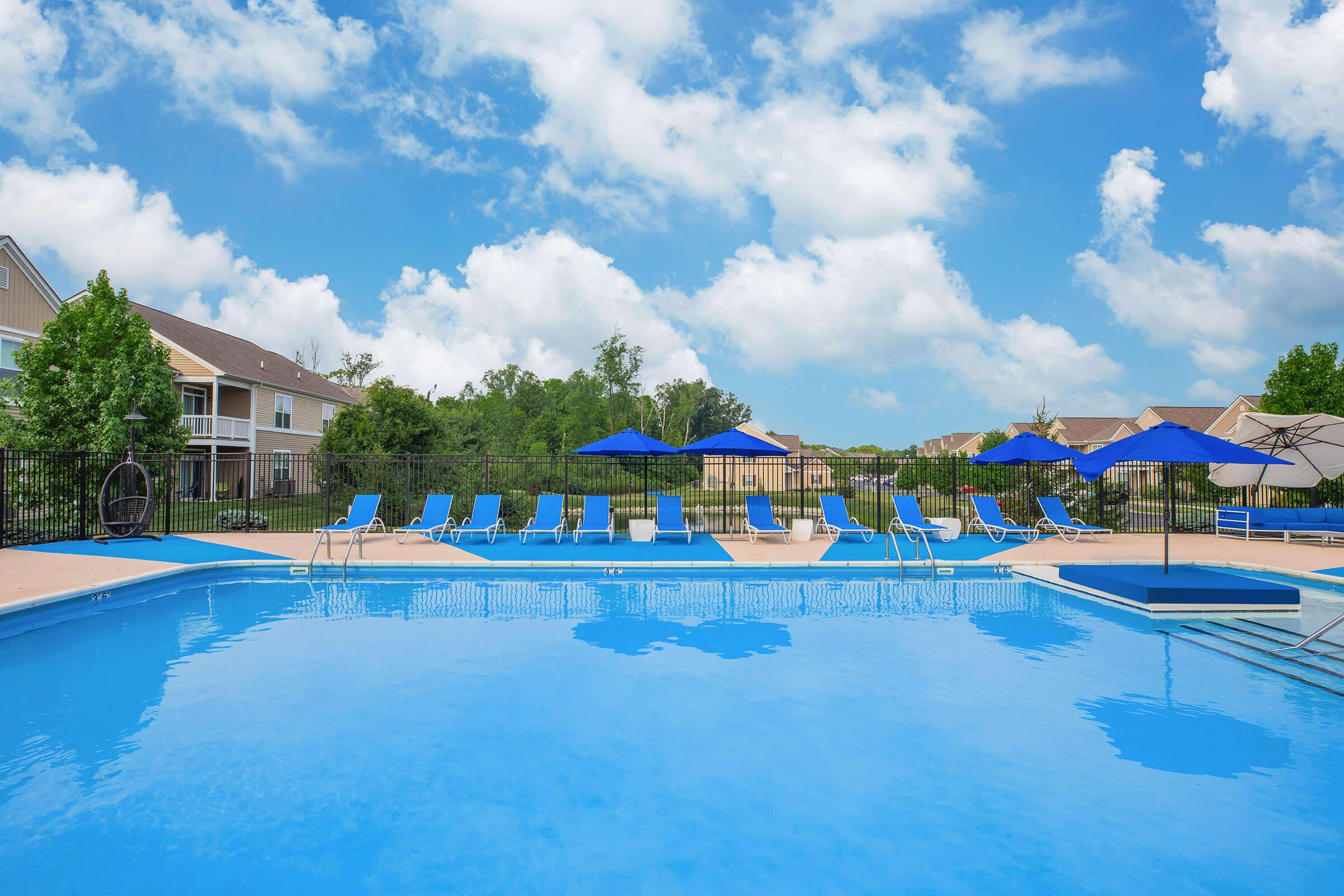 A bright blue swimming pool surrounded by lounge chairs and umbrellas under a partly cloudy sky. Trees and residential buildings are visible in the background.