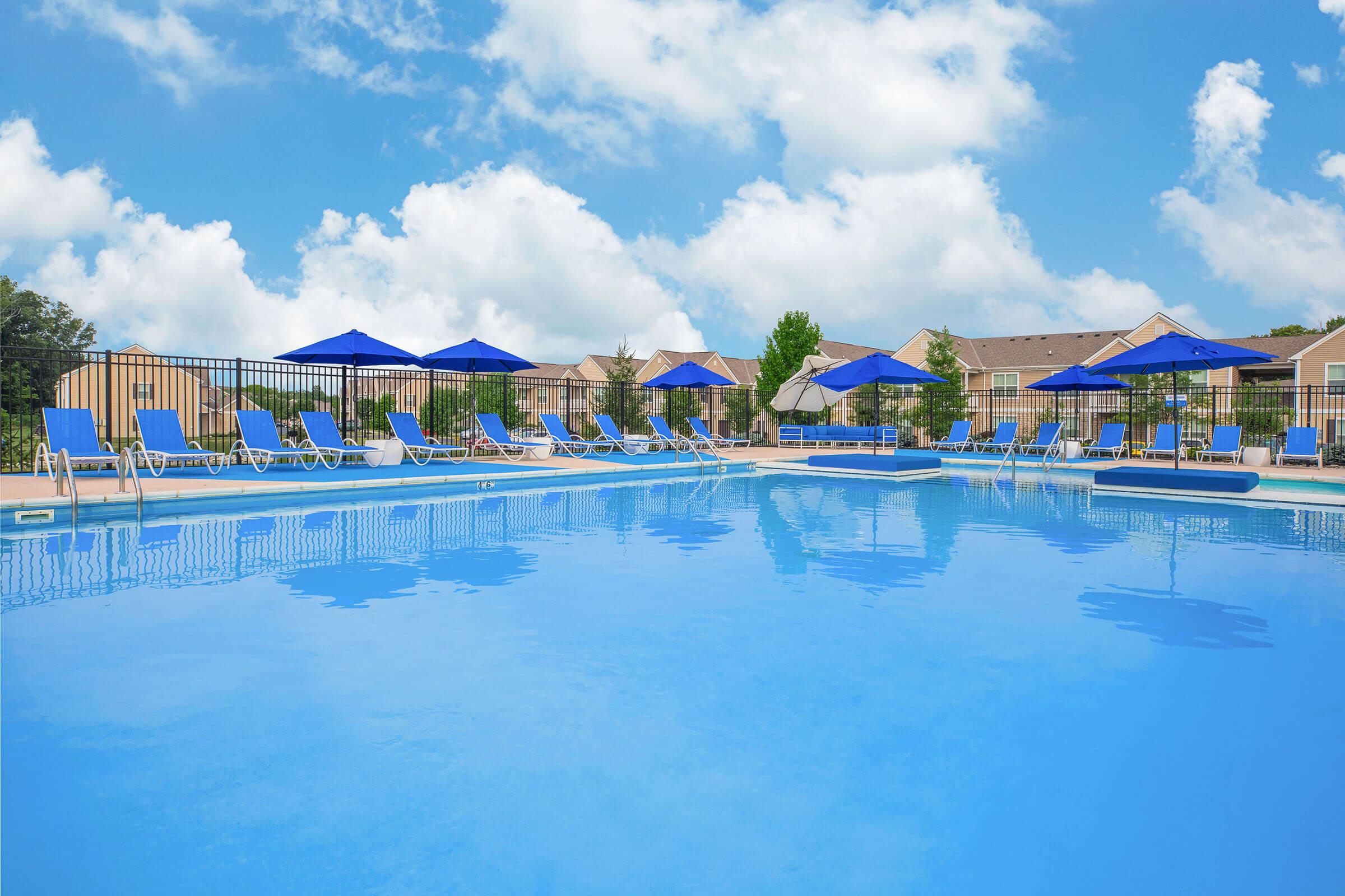 A sunny pool scene featuring a clear blue pool with lounge chairs and umbrellas around the edge, set against a backdrop of fluffy white clouds and greenery.