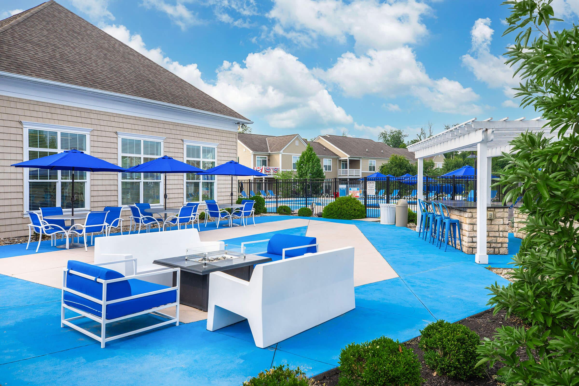 A well-maintained outdoor pool area featuring blue and white lounge furniture, umbrellas, and a seating area with a modern fire pit. Surrounding greenery and a blue sky with clouds enhance the inviting atmosphere.