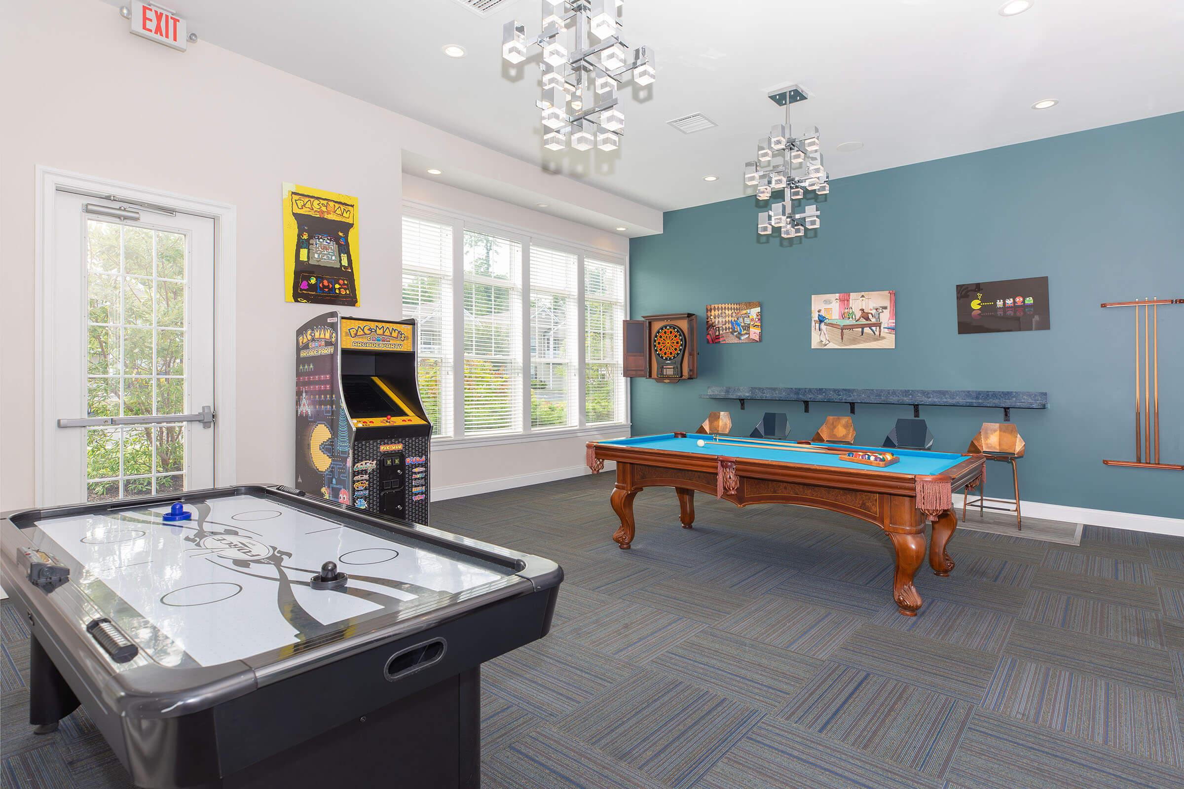 A recreational room featuring a pool table, an air hockey table, and an arcade game. The walls are adorned with colorful artwork, and there are several chairs around the pool table. Large windows allow natural light to enter the space.