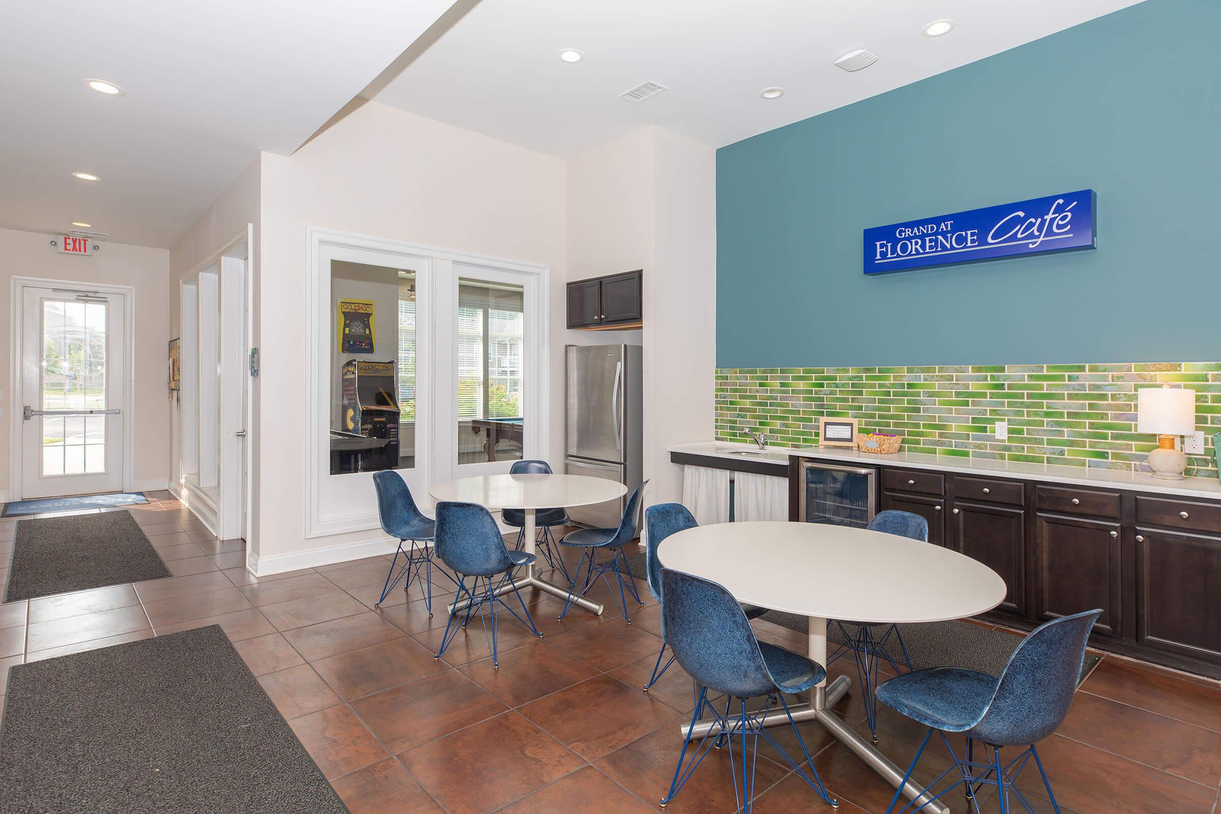 A modern café area with two round tables and blue chairs, featuring a colorful green tile backsplash and a sign that says "Grand of Florence Café." The space has large windows allowing natural light in, with a stainless steel refrigerator and other kitchen appliances visible.