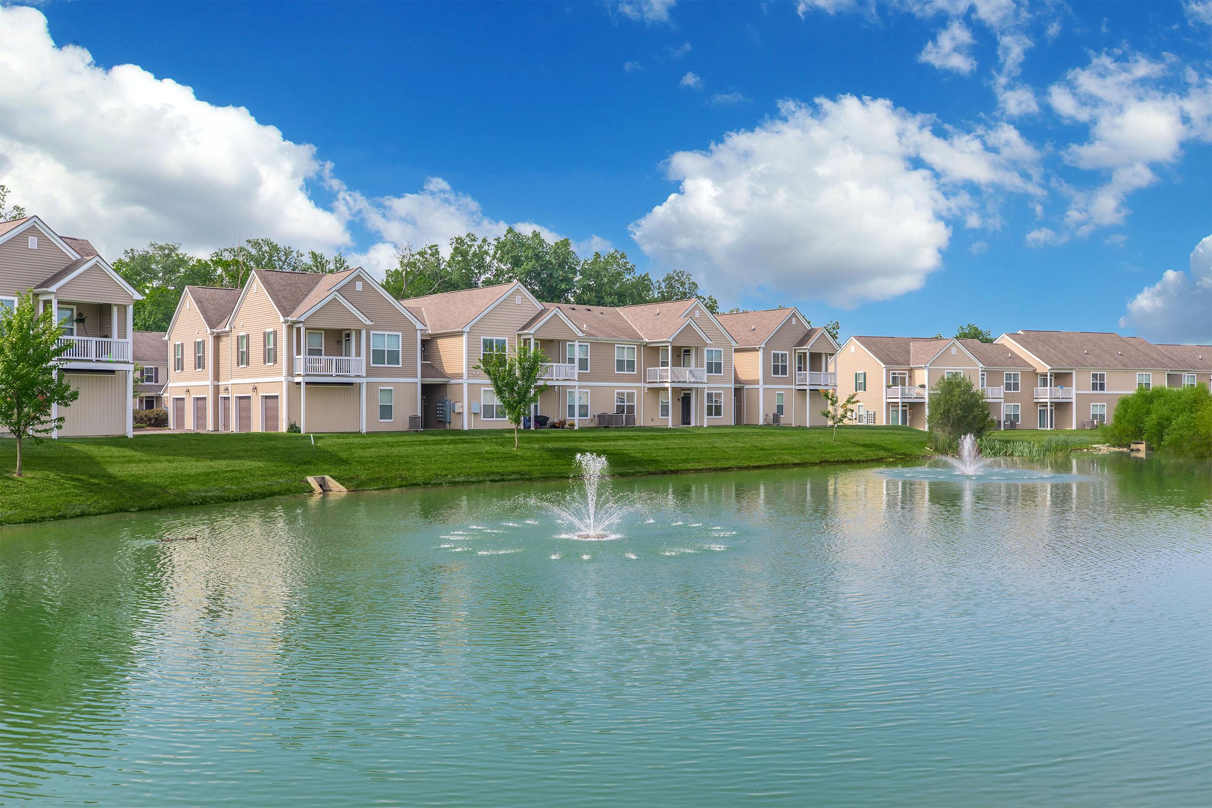 A serene residential area featuring two-story apartment buildings beside a calm pond. The pond has decorative fountains, and the landscape includes green lawns and trees under a bright blue sky with fluffy white clouds.