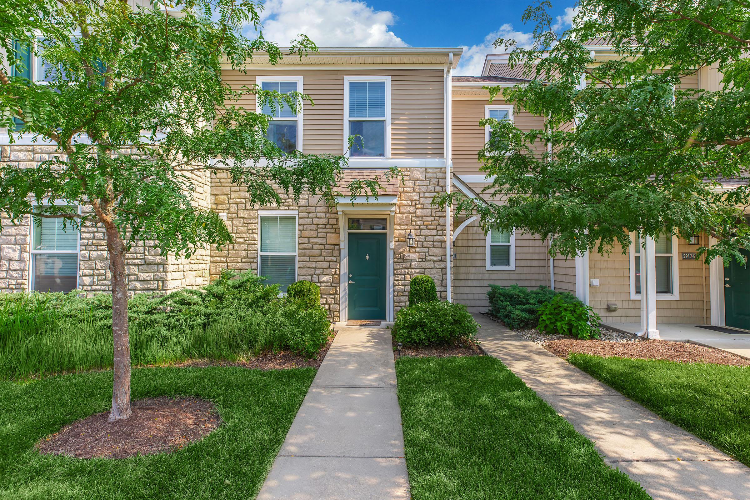 A well-maintained two-story townhouse with a stone and siding exterior. The entrance features a teal door, surrounded by lush greenery, including small trees and shrubs. A concrete walkway leads to the front door, and the sky is clear with a few clouds, indicating a sunny day.