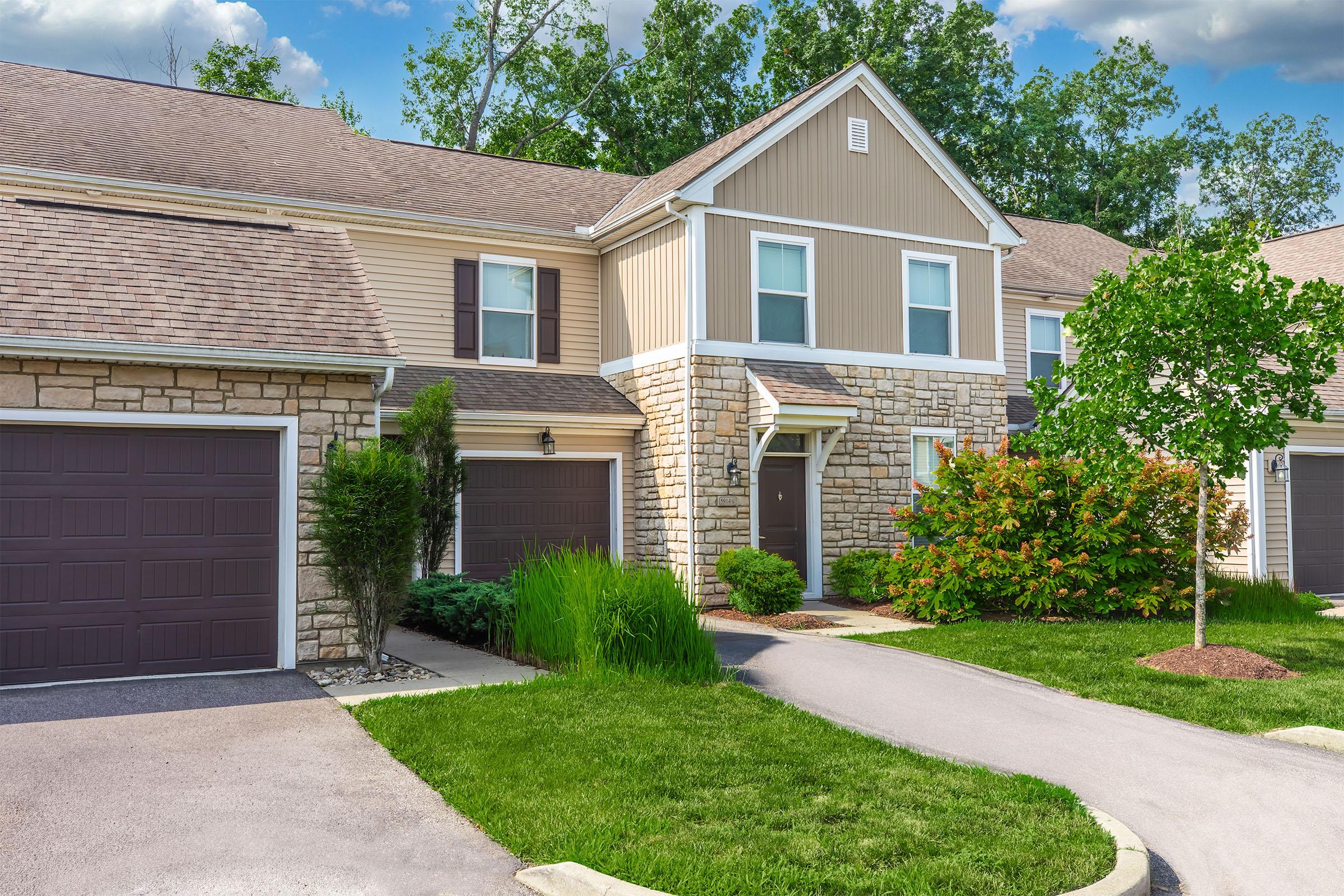 A residential townhouse with a stone and siding exterior, featuring a paved driveway, green lawn, and well-maintained shrubs. The building has two garages with brown doors and a front entrance surrounded by greenery under a clear blue sky.