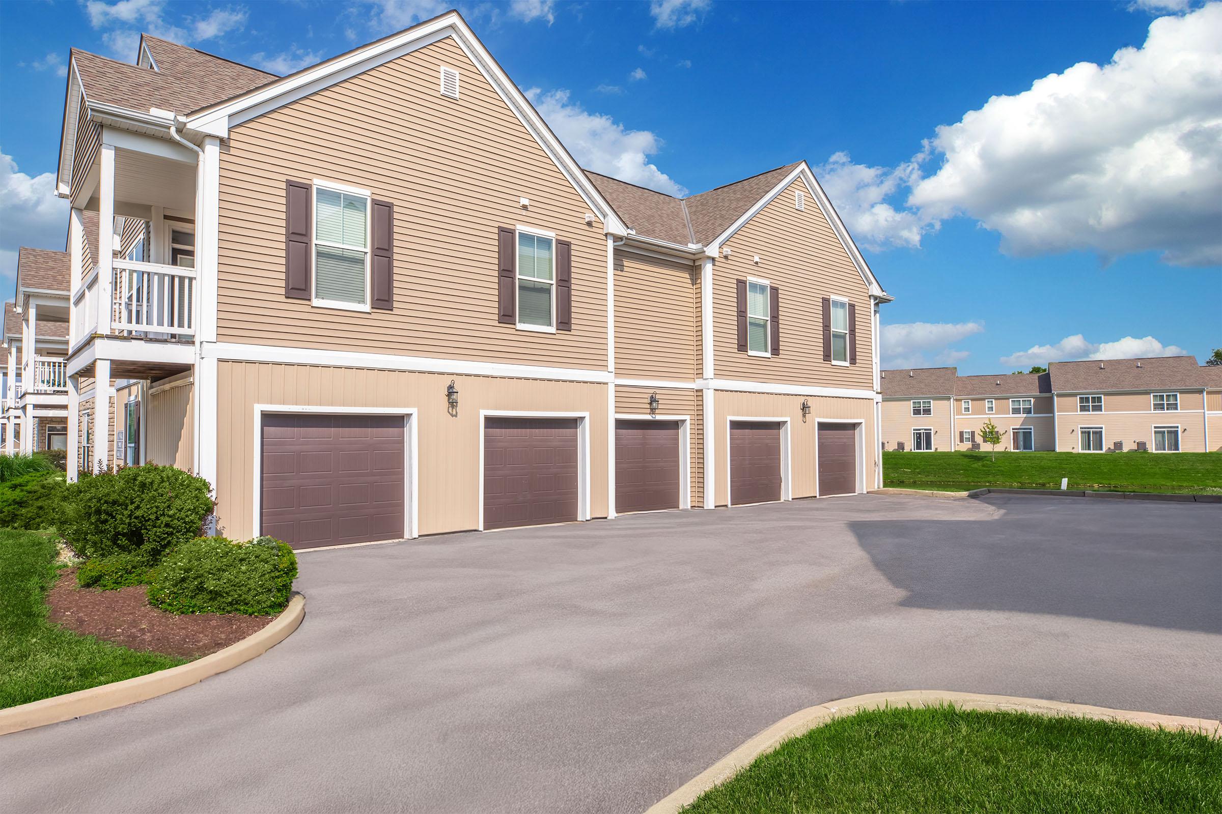 A modern residential building featuring a light brown exterior with multiple garage doors. The setting includes a paved driveway and well-maintained green lawns. In the background, additional similar buildings can be seen under a clear blue sky with a few clouds.