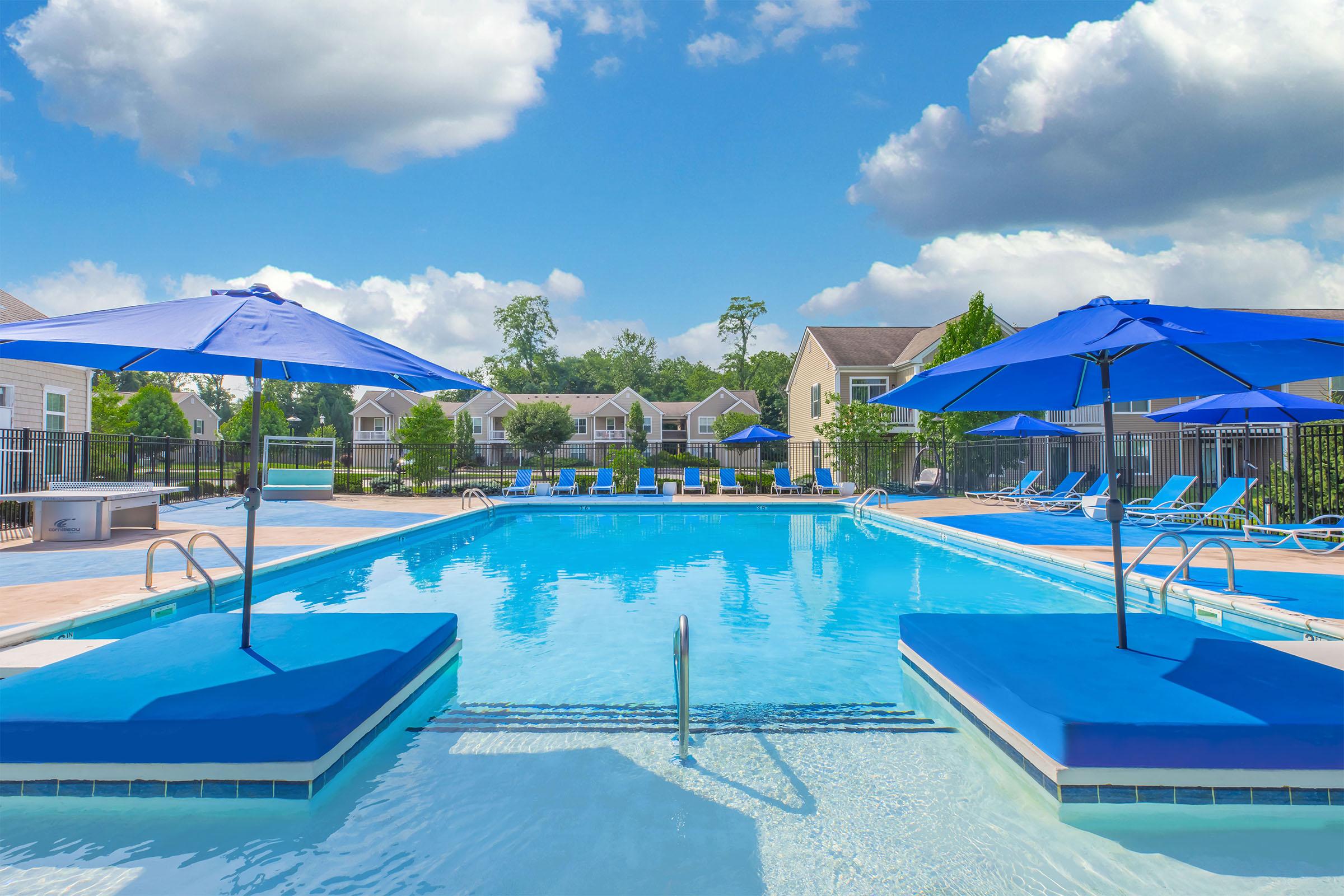 A sunny outdoor pool area featuring crystal-clear water, blue umbrellas providing shade, and lounge chairs on the deck. Surrounding the pool are well-maintained residential buildings and greenery, creating a relaxing environment. The sky is bright with fluffy clouds.