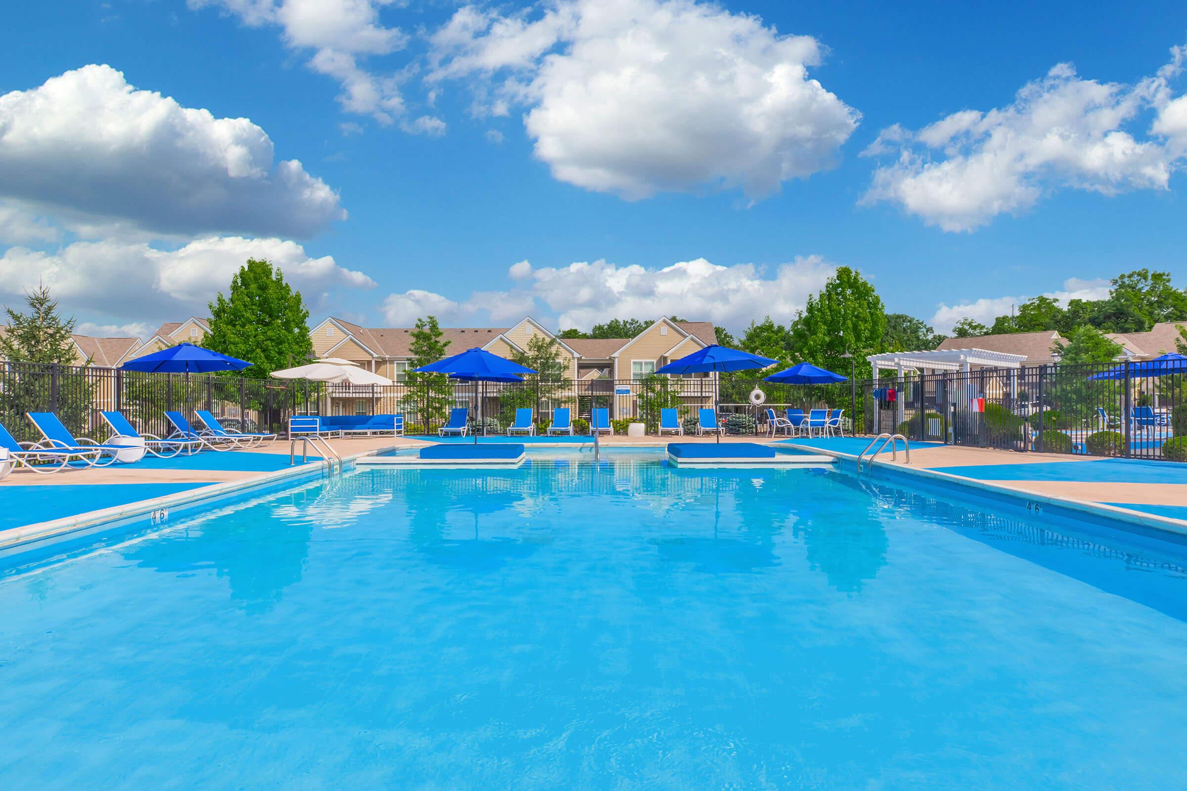 View of a bright blue swimming pool surrounded by lounge chairs and umbrellas. The scene features a clear sky with fluffy clouds and residential buildings in the background. Lush trees and well-maintained landscaping enhance the outdoor setting, creating a relaxing atmosphere.