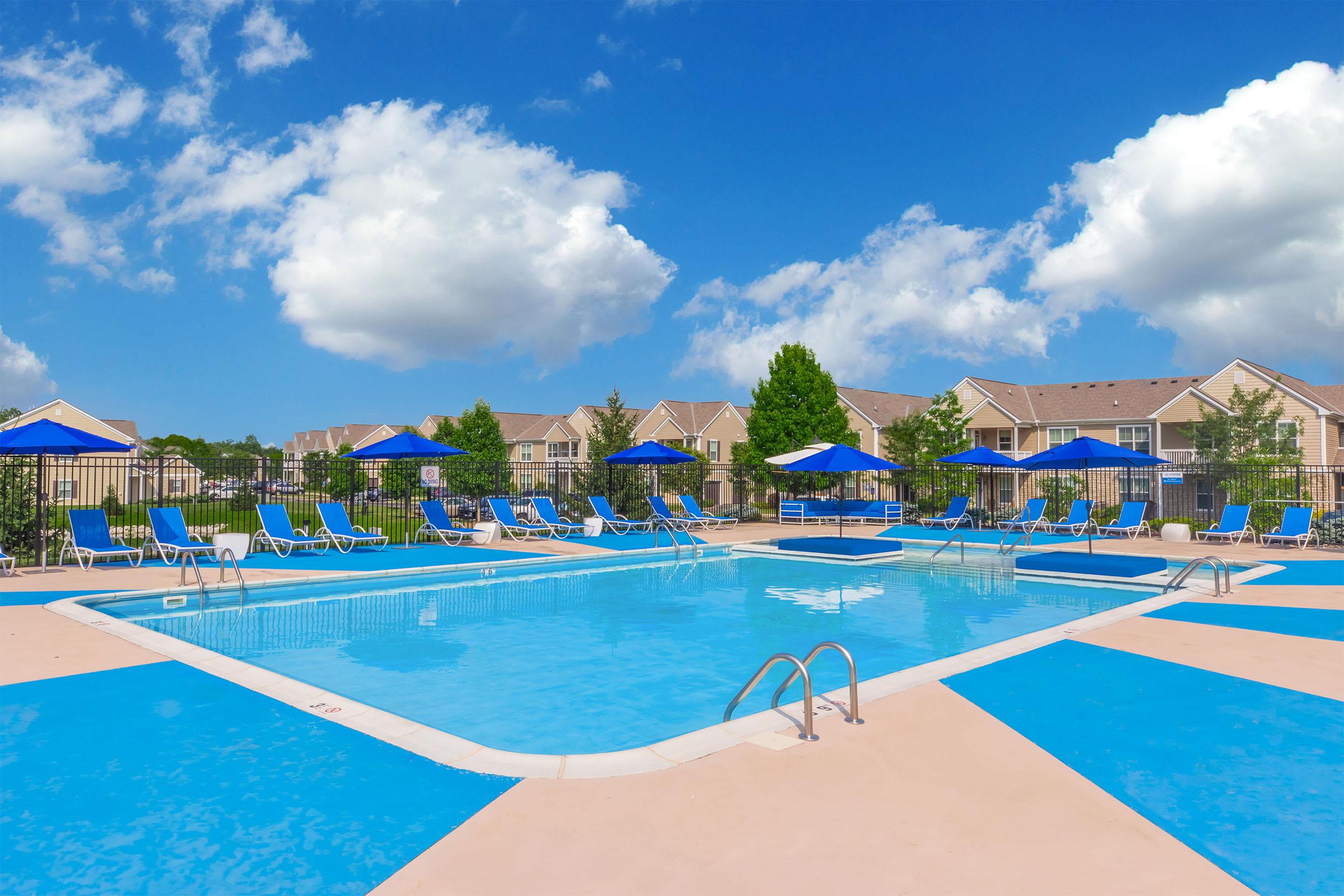 A bright outdoor swimming pool surrounded by lounge chairs and blue umbrellas, with a clear blue sky and fluffy white clouds in the background. Nearby, there are landscaped areas with trees and residential buildings visible. The scene conveys a relaxing and inviting atmosphere.
