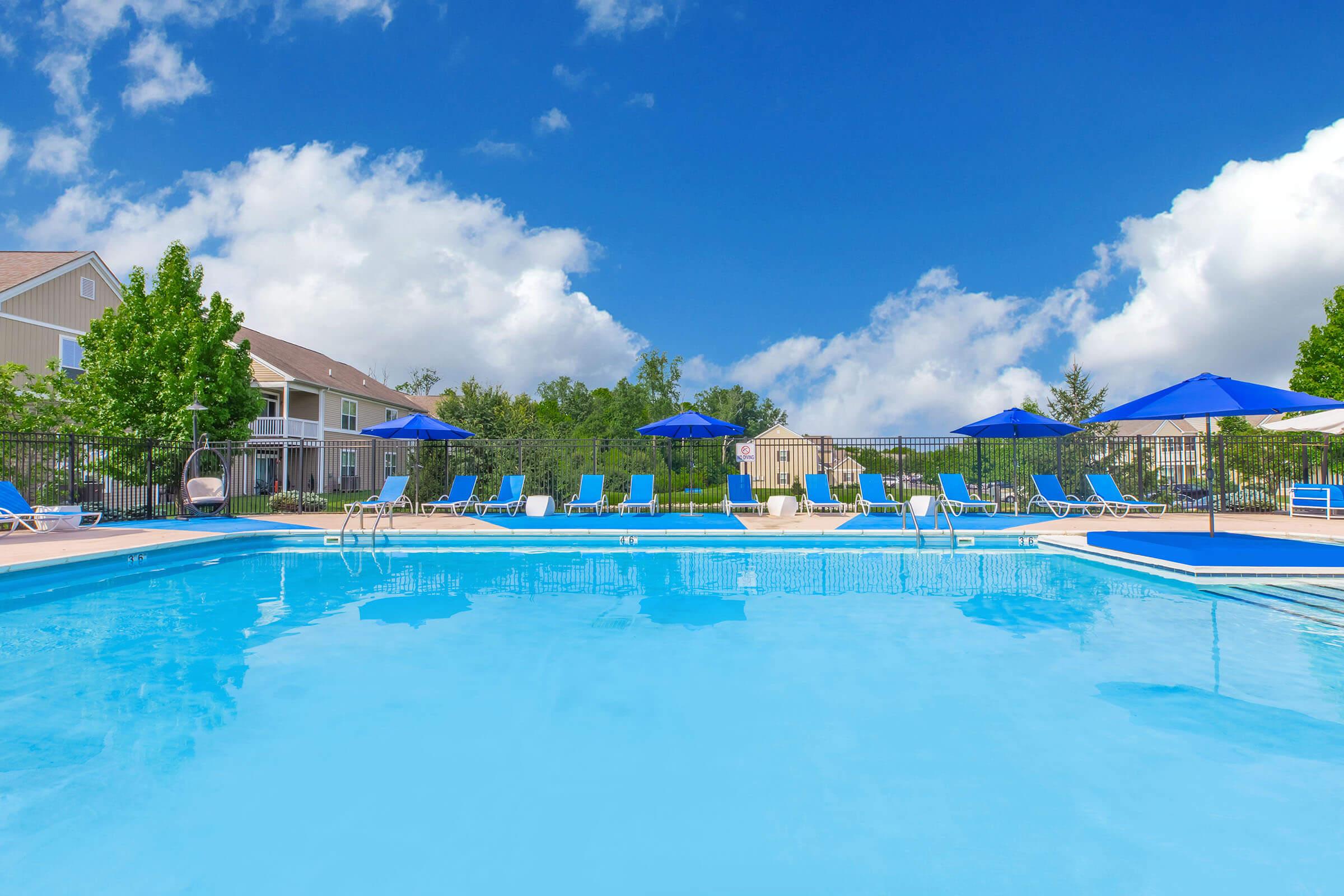 A clear blue swimming pool surrounded by lounge chairs and umbrellas, set against a backdrop of green trees and a blue sky with fluffy white clouds. The pool area features neatly arranged lounge chairs, creating a relaxing atmosphere for visitors.