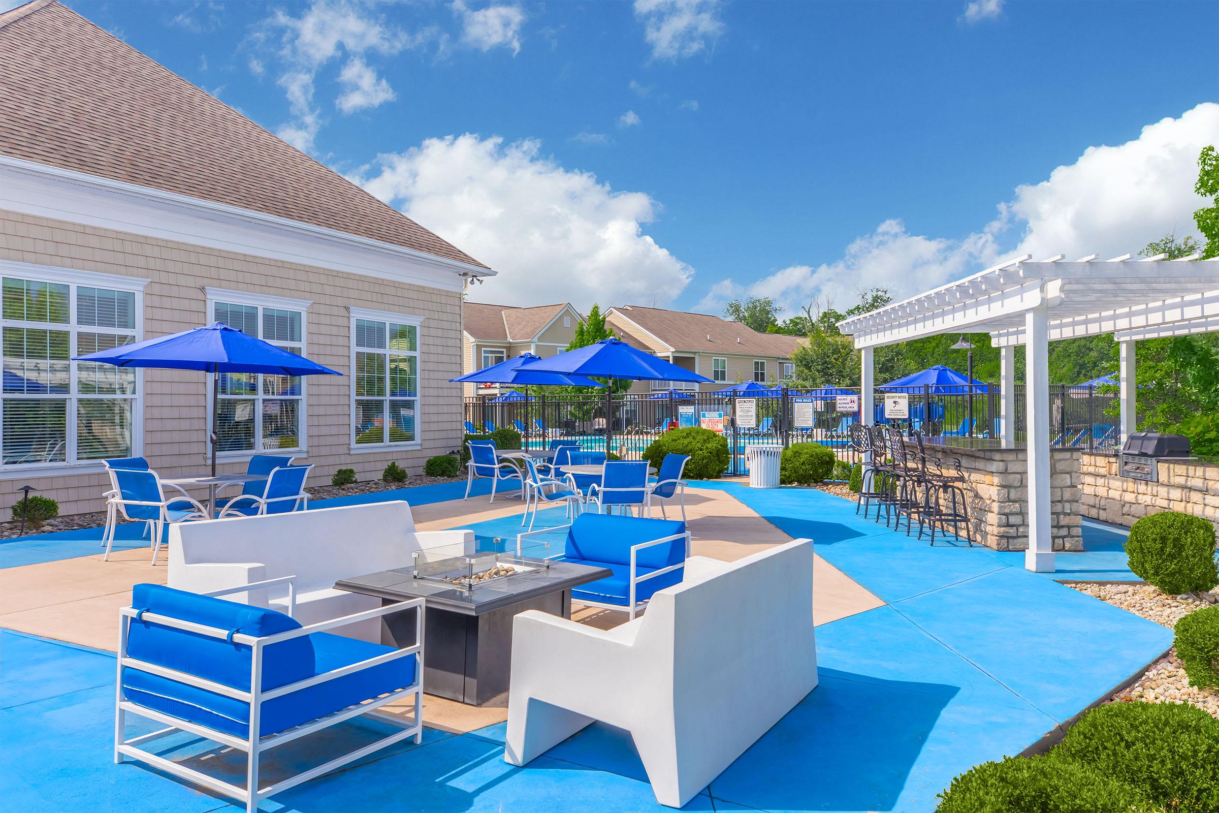 A sunny outdoor patio area featuring blue and white lounge chairs, a fire pit table, and several blue umbrellas. Surrounding greenery and a building with large windows complete the scene, offering a relaxing space for socializing and enjoying the weather.