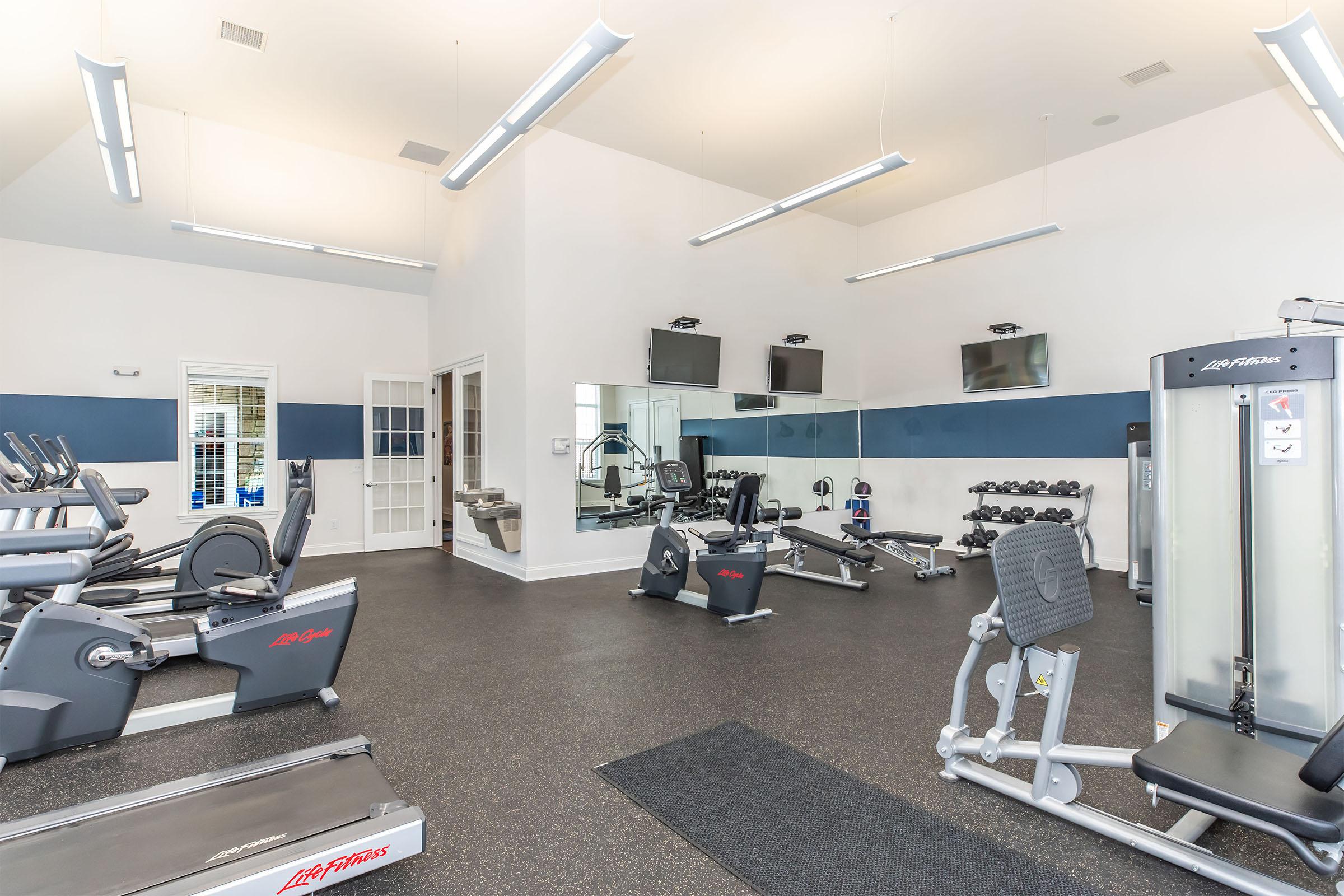A bright and spacious gym area featuring various exercise equipment, including treadmills, stationary bikes, weight machines, and free weights. The space is well-lit with overhead lights and includes multiple mirrors and televisions mounted on the walls.