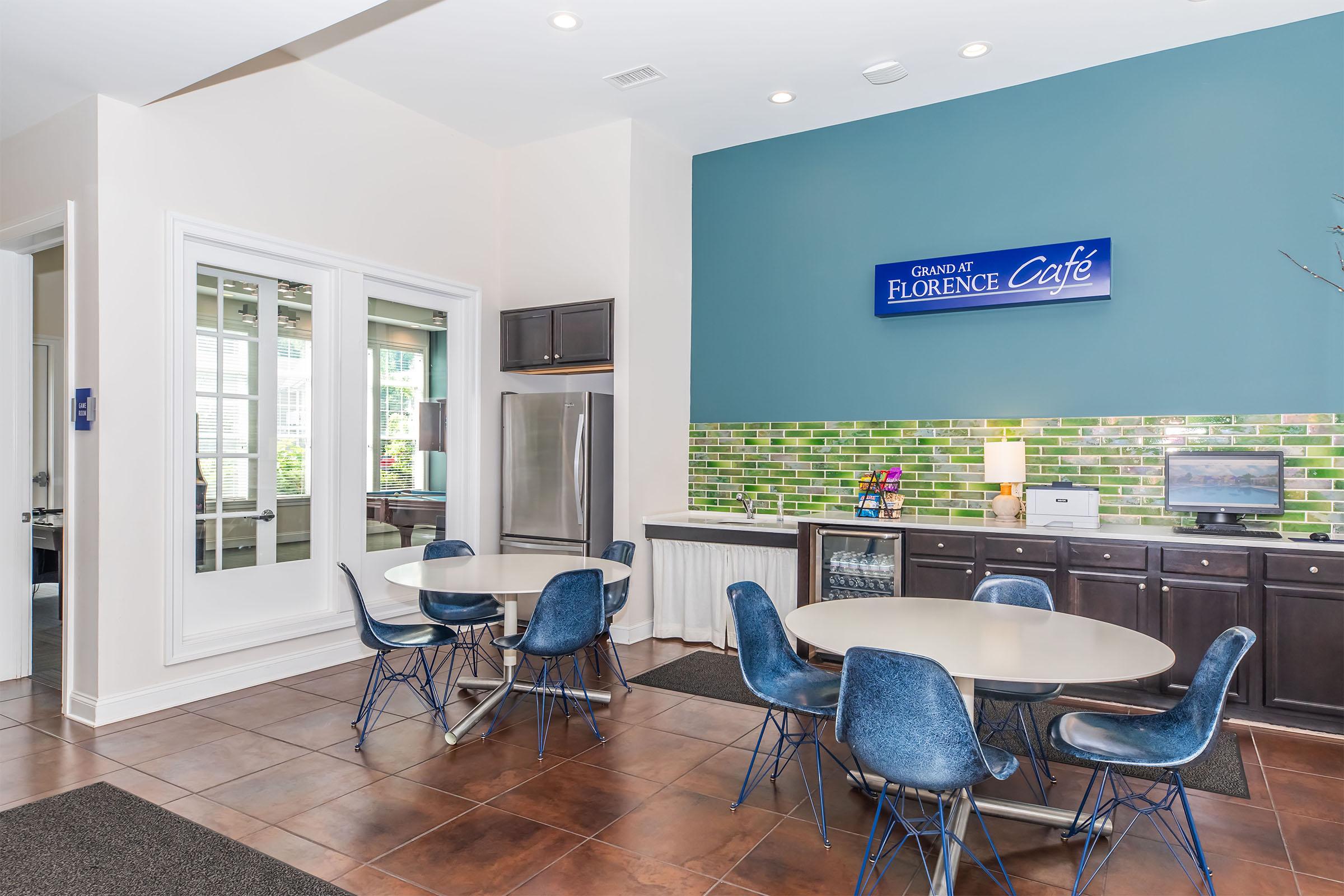 Bright café area featuring modern decor with green tiled backsplash, stainless steel appliances, and a bank of windows allowing natural light. There are several round tables with blue chairs and a countertop with coffee-making facilities, creating a welcoming atmosphere for guests to relax.