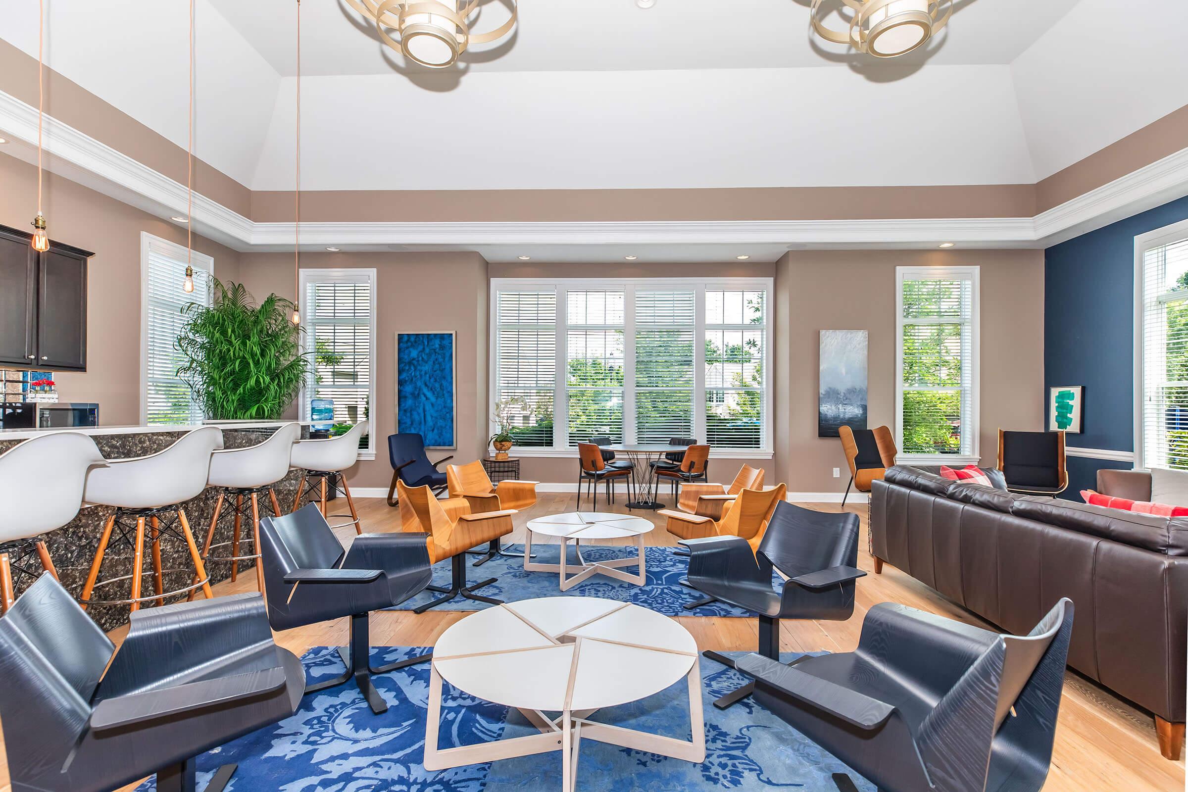 A spacious, modern living area featuring stylish furniture, including mid-century chairs and a sleek coffee table. Large windows let in natural light, showcasing green views outside. The room has a cozy atmosphere with neutral walls, decorative art, and plants, creating an inviting space for relaxation and socializing.