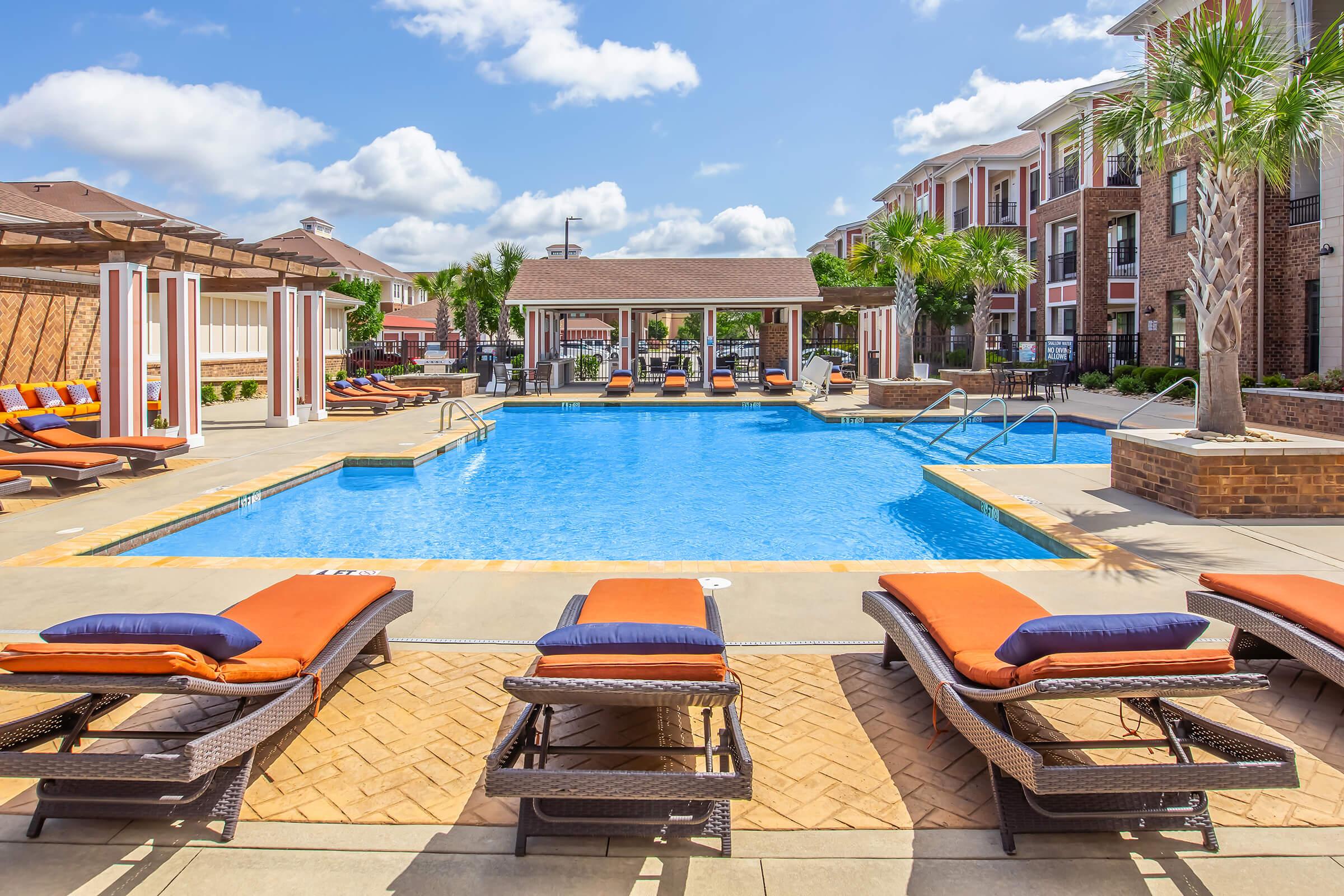 A spacious outdoor pool area featuring sun loungers with orange cushions, surrounded by well-maintained landscaping. In the background, there are modern apartment buildings and a shaded cabana offering seating. The sky is clear with a few clouds, creating a bright and inviting atmosphere.