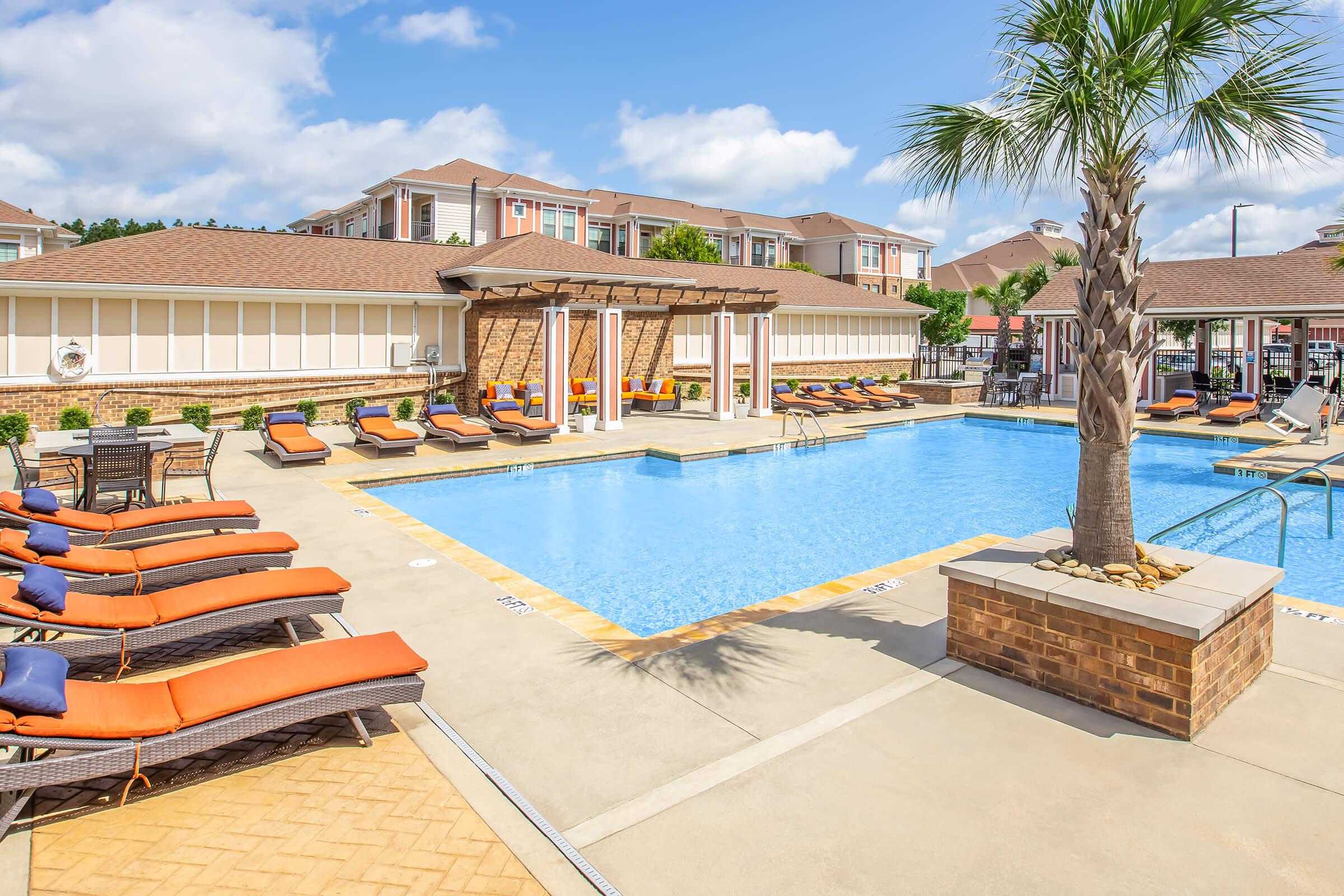 A sunny outdoor pool area featuring a large swimming pool surrounded by lounge chairs with orange cushions. Palm trees add a tropical touch, and the pool is bordered by a decorative stone wall. The background includes residential buildings under a partly cloudy sky, creating a relaxing atmosphere.