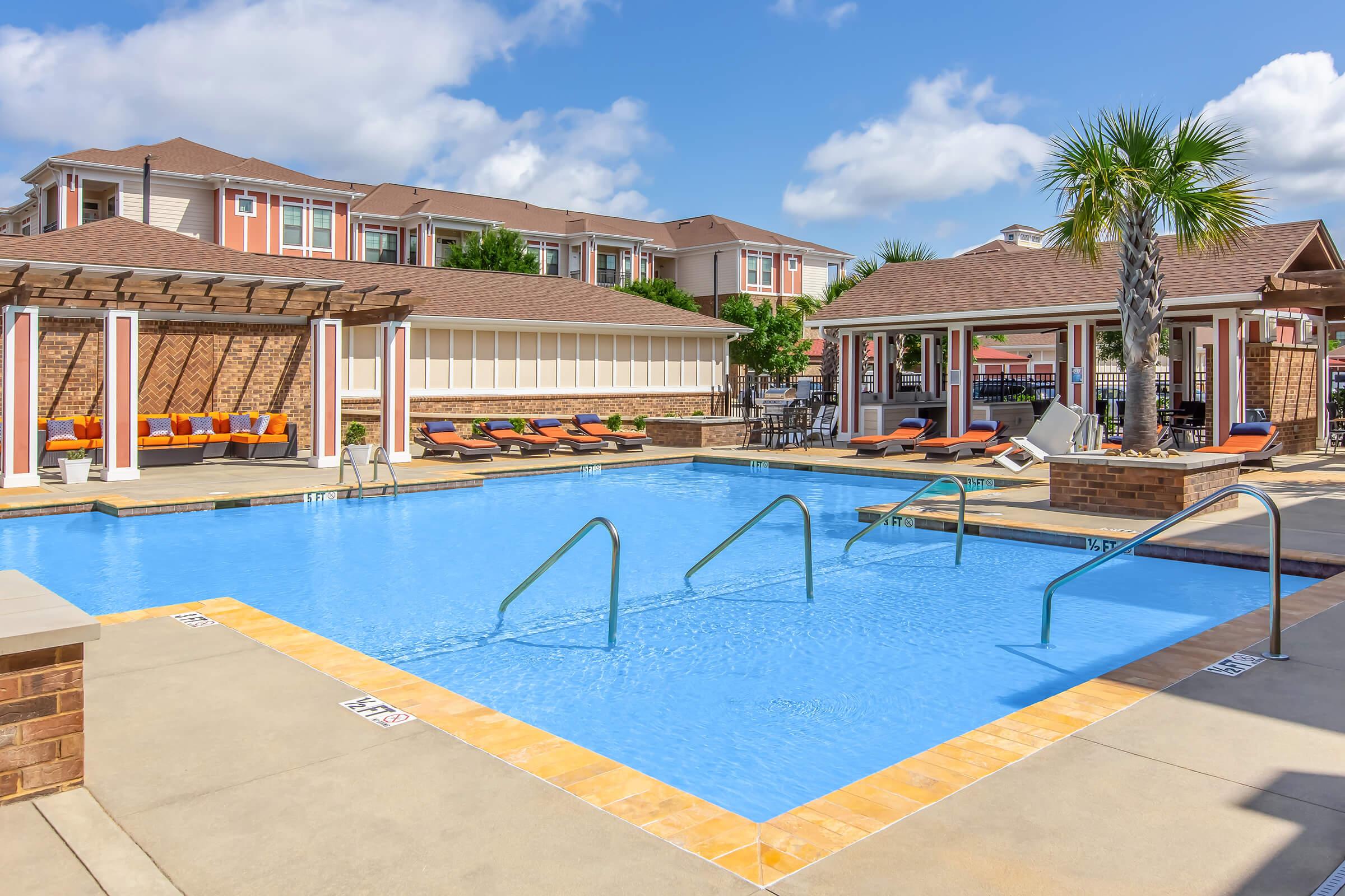 A bright outdoor swimming pool surrounded by lounge chairs and palm trees. Two shaded areas with pergolas are visible, along with sunbathers relaxing on the deck. Modern apartment buildings provide a backdrop under a clear blue sky. The scene conveys a warm and inviting atmosphere for leisure.