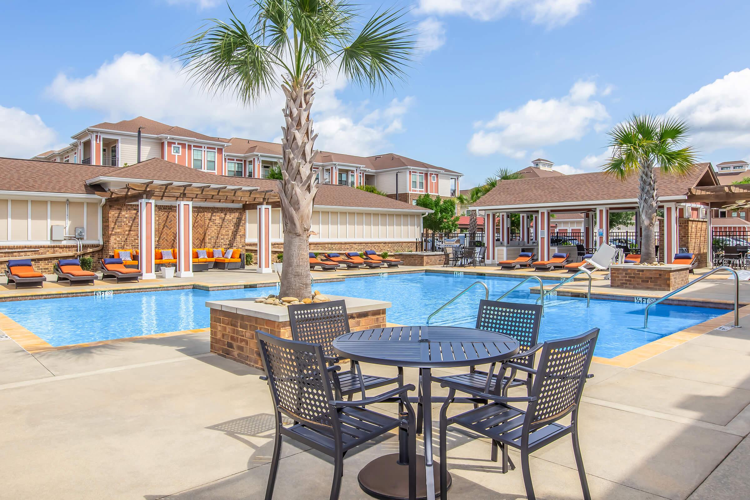 A sunny pool area featuring a large swimming pool surrounded by lounge chairs. There are palm trees and a patio with a table and chairs in the foreground. In the background, multiple residential buildings are visible, creating a relaxing atmosphere.