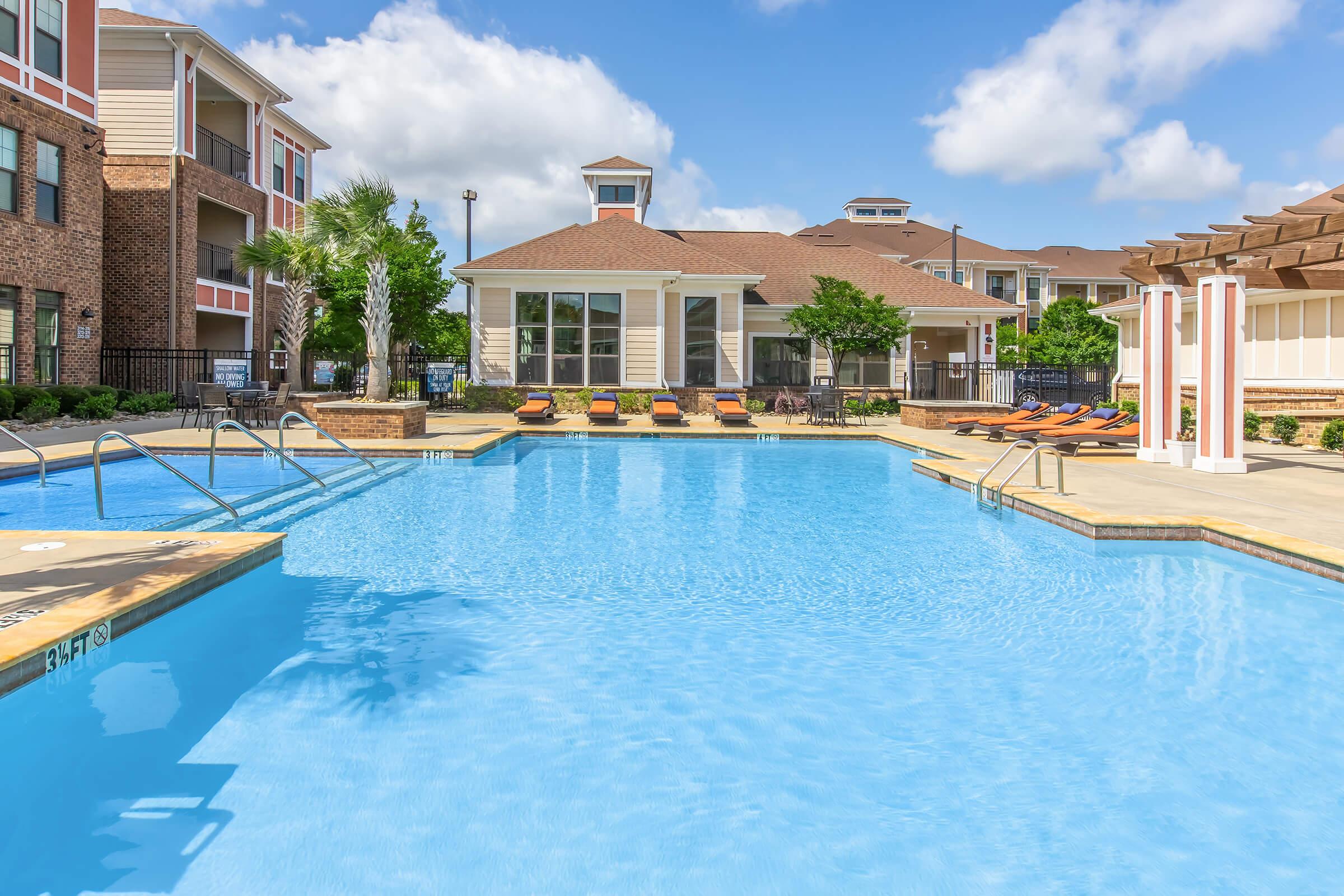 A clear blue swimming pool surrounded by lounge chairs and palm trees, with a clubhouse in the background under a sunny sky. The area features well-maintained landscaping and a relaxing atmosphere, perfect for leisure and recreation.