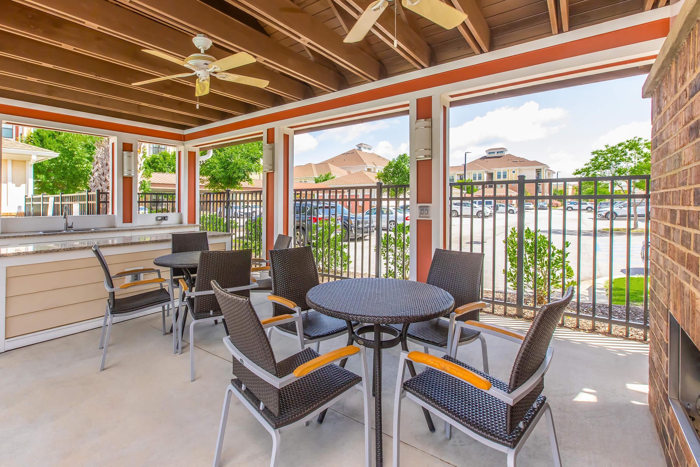 An outdoor seating area with a black table and six chairs, shaded by a roof with ceiling fans. Nearby, there's a bar area with a countertop. The background shows a gated parking lot and green trees under a clear blue sky.