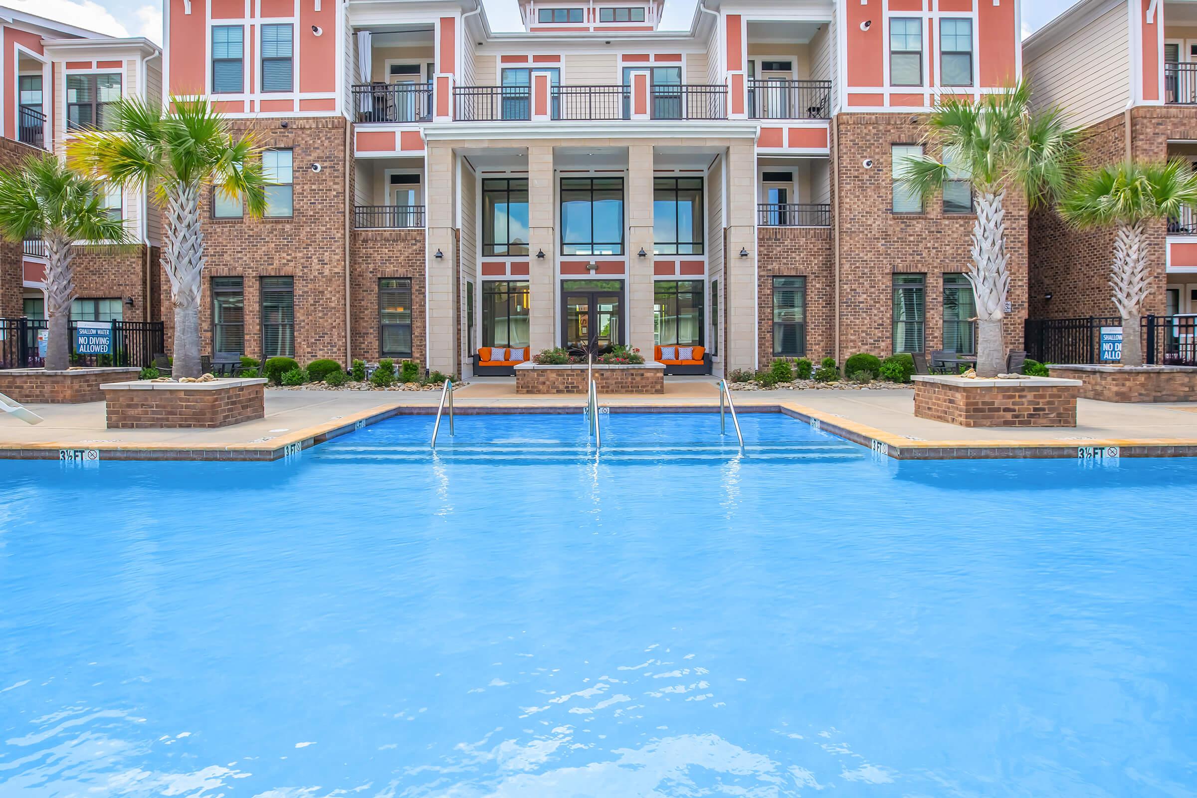 A clear blue swimming pool with steps leading out of it, located in front of a large brick and colorful apartment building. The pool area is surrounded by palm trees and landscaped greenery, with several lounge chairs positioned around the edges. Large windows in the building reflect the pool's tranquil water.