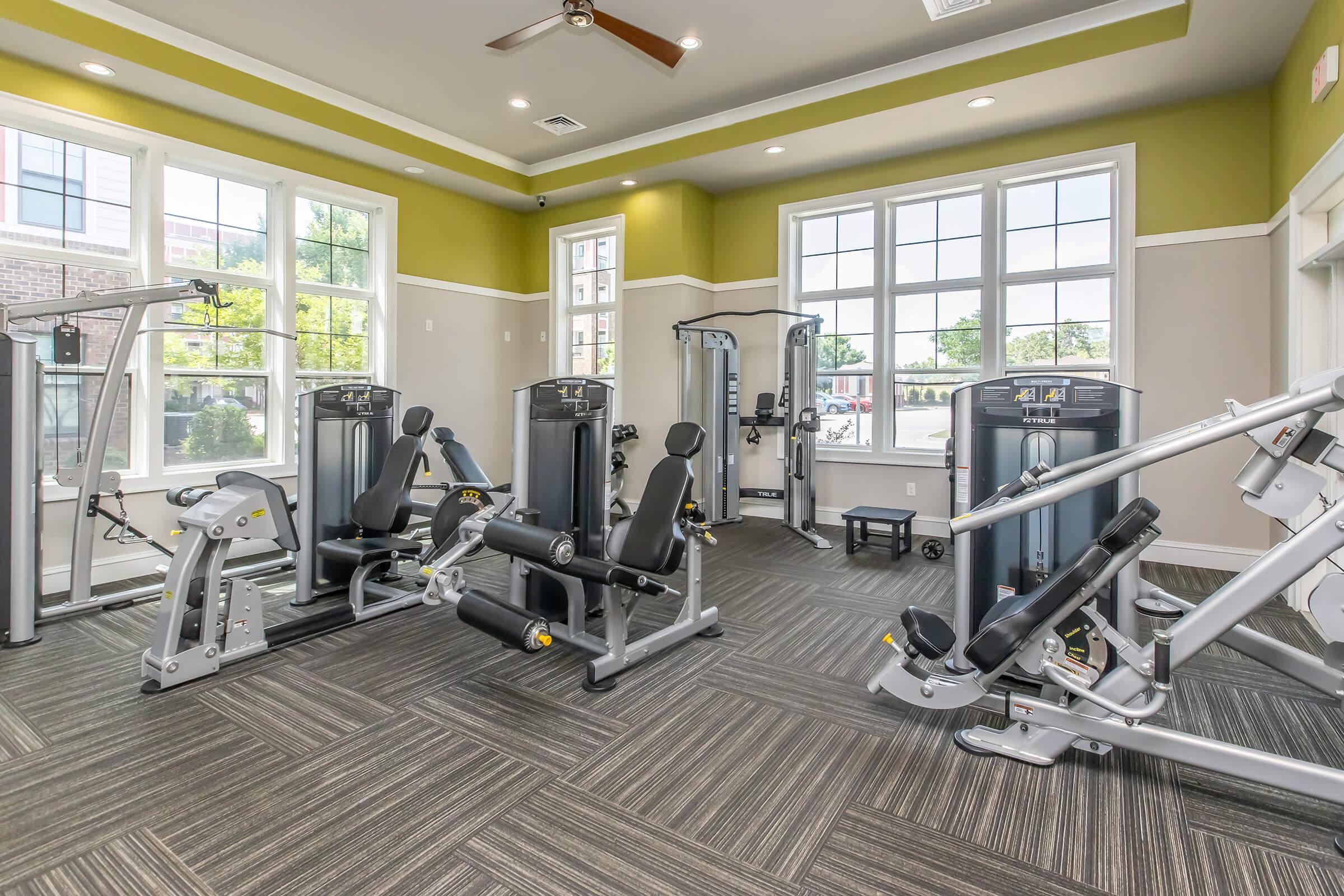 A modern gym interior featuring various exercise machines, including weight-training equipment, set against a backdrop of large windows allowing natural light. The space has neutral-colored walls with green accents and a polished floor, creating a bright and inviting atmosphere for workouts.