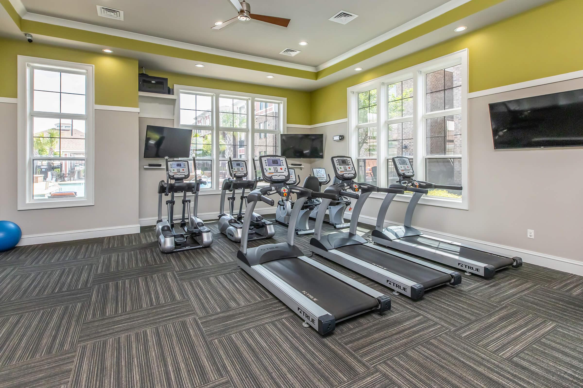 A modern fitness room featuring several treadmills arranged in a row, equipped with screens. The space has large windows allowing natural light, and is finished with a neutral color palette and a patterned dark floor. Two televisions are mounted on the walls, enhancing the workout environment.
