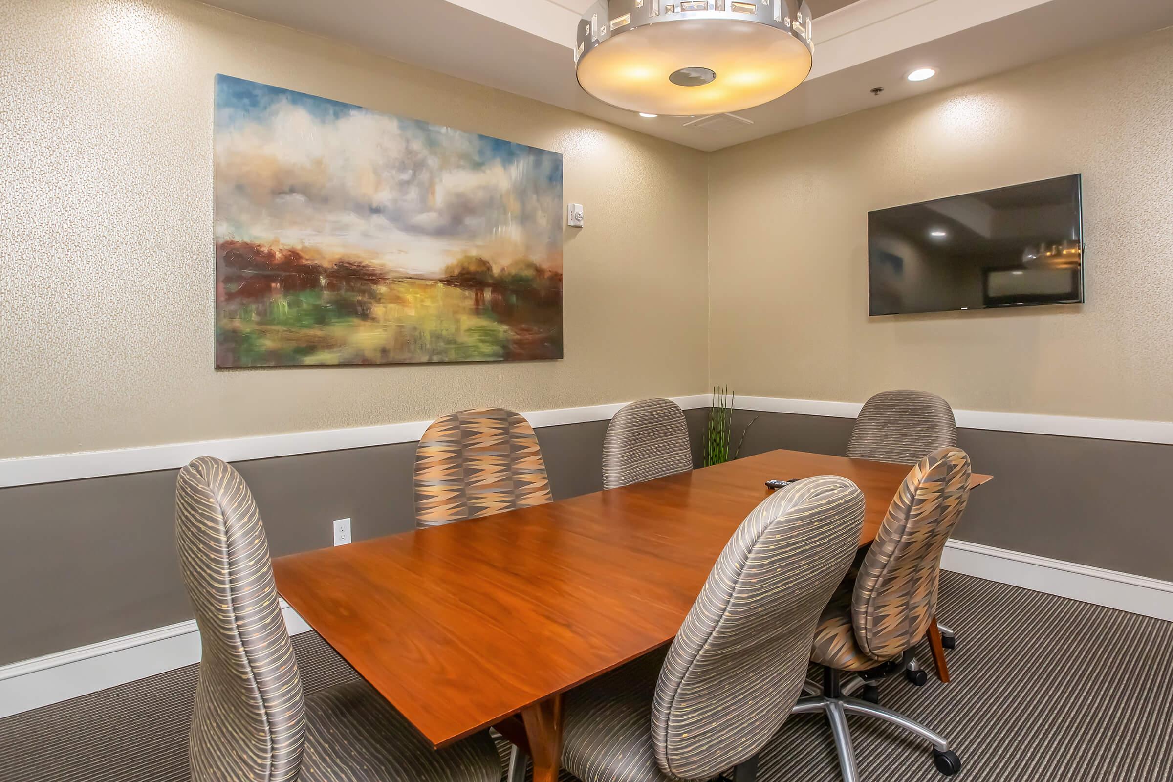 A modern conference room featuring a wooden table surrounded by ergonomic chairs. The walls have a textured design, with a large abstract painting hanging at one end. A flat-screen TV is mounted on the opposite wall, and there is a small plant in the corner, adding a touch of greenery to the space.