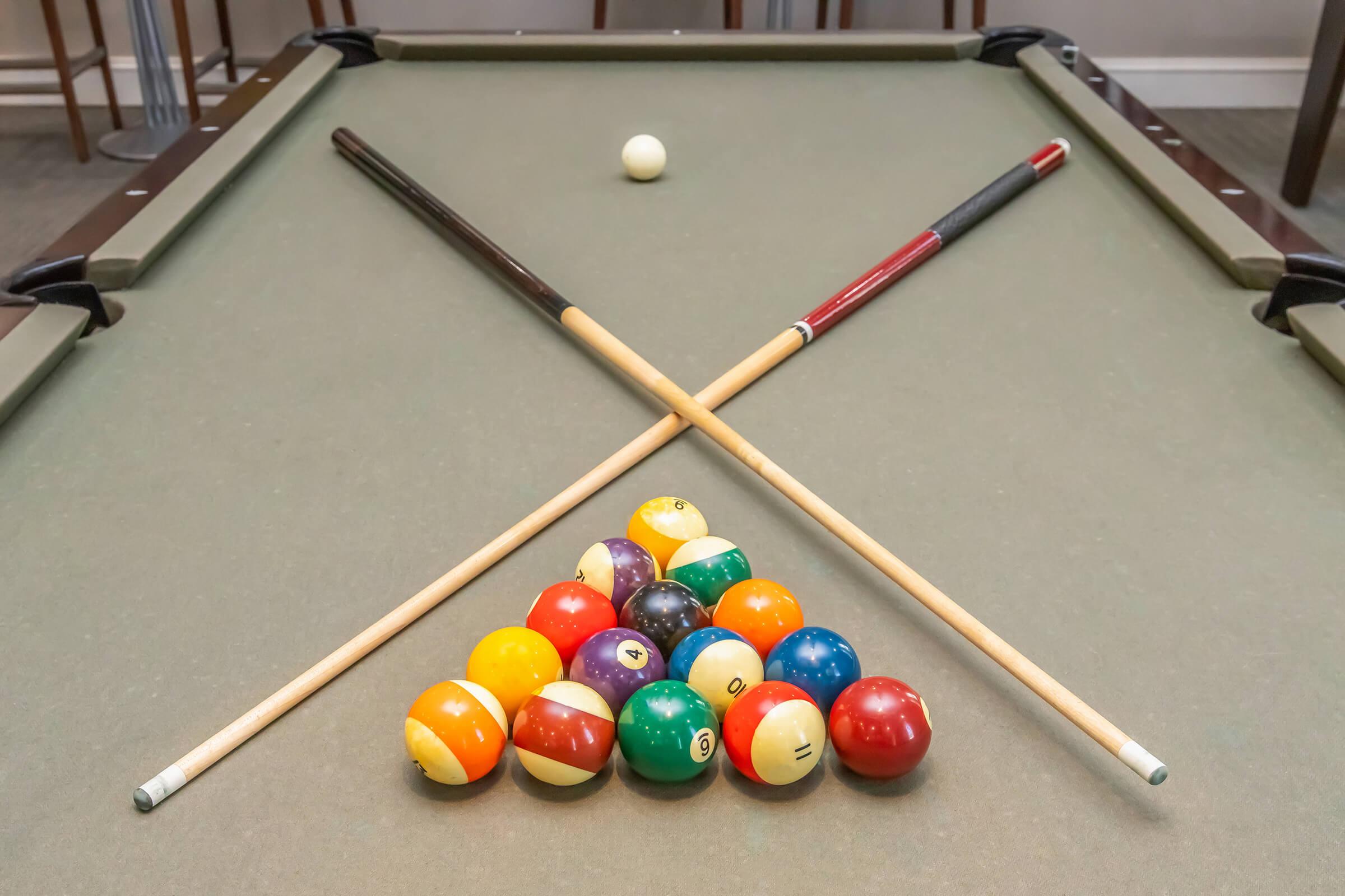 A pool table with a triangle rack of billiard balls arranged at the bottom. Two pool cues are crossed over the table. A single cue ball is positioned above the rack, ready for the game to begin. The background shows a neutral-toned space with bar-style seating.
