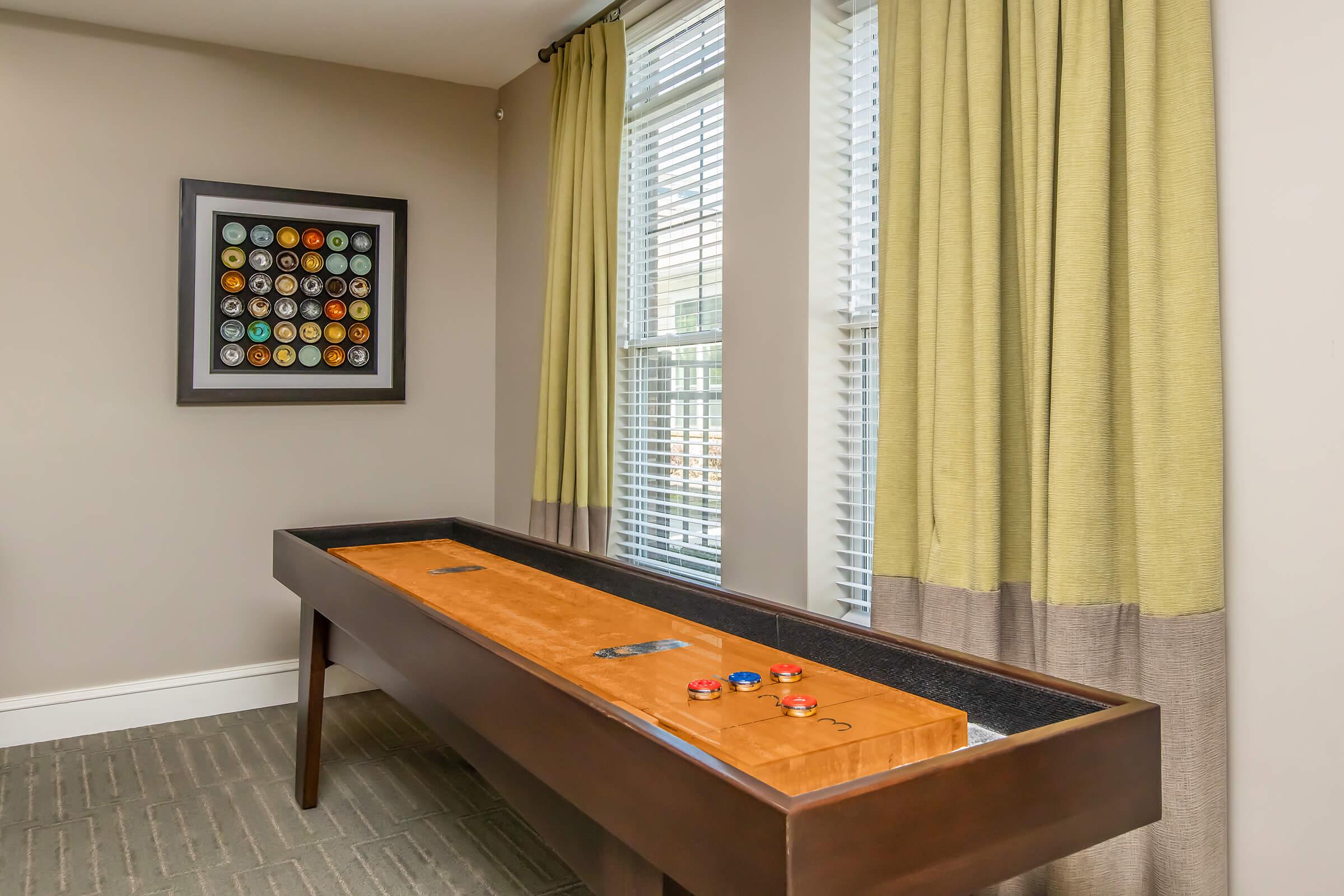 A shuffleboard table with a polished wooden surface is positioned against a wall in a well-lit room. There are colored pucks on the table, and large windows with light green curtains add to the ambiance. A framed circular artwork is displayed on the wall, complementing the modern decor.