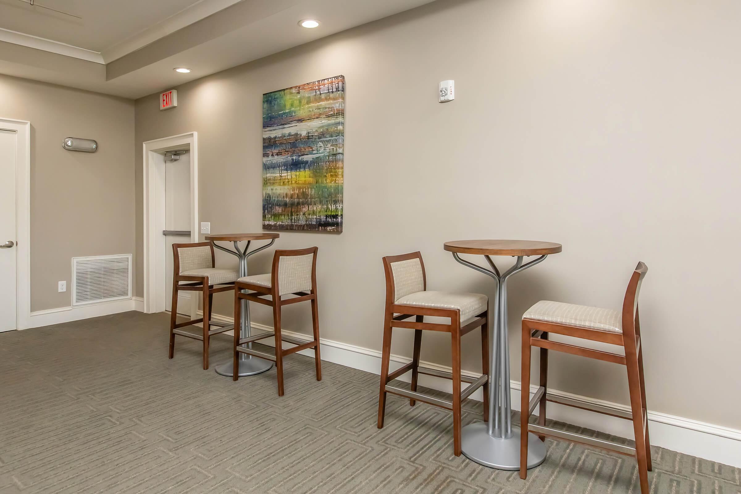 A modern lounge area featuring two tall tables with stools, set against a light beige wall. The decor includes a colorful abstract painting above the tables and a door leading to another room. The floor is covered with a textured carpet, adding warmth to the space.