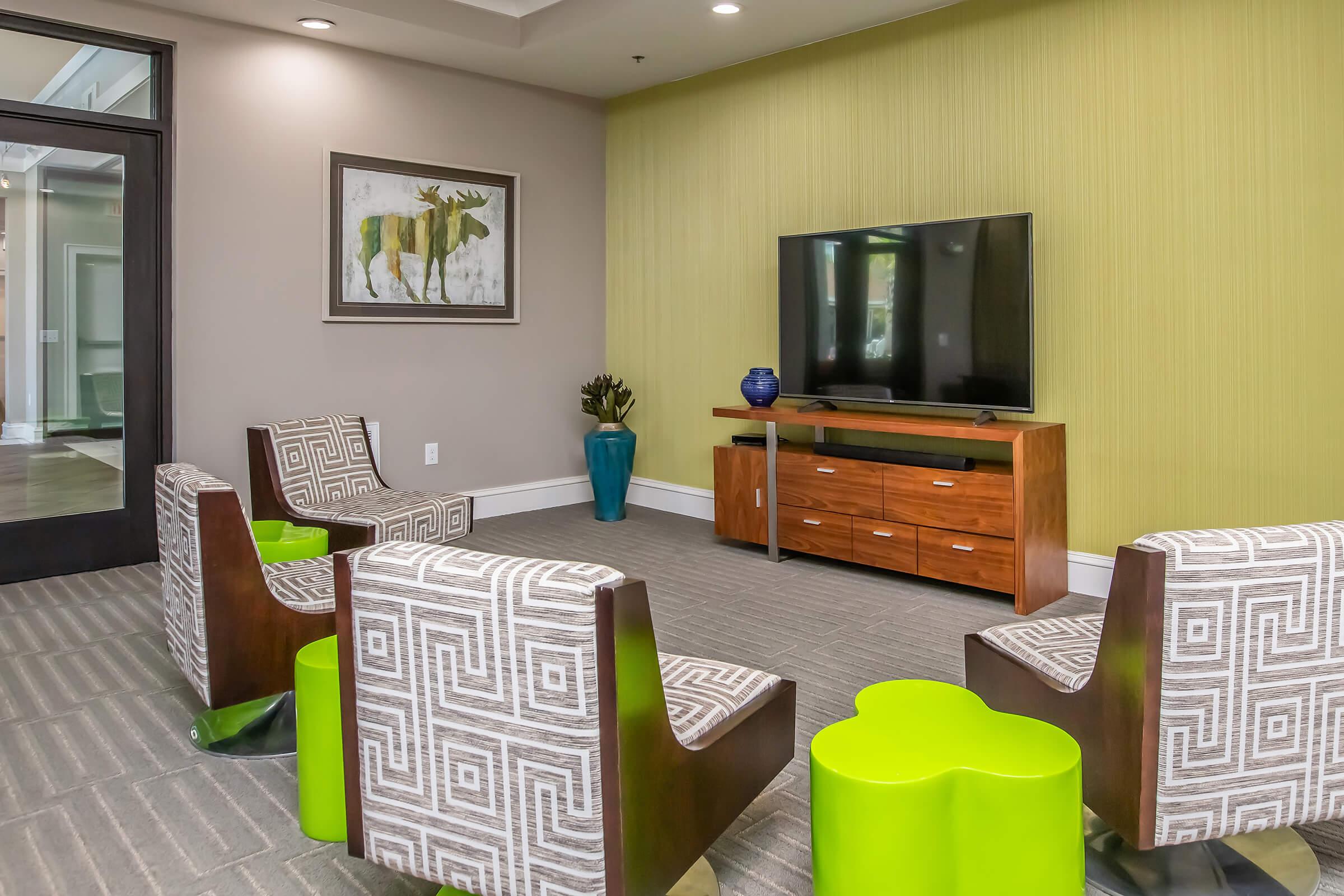 A modern lounge area featuring two patterned armchairs and a green and wooden coffee table. A television is mounted on the wall above a wooden TV stand. There are decorative elements including a blue vase and artwork of a moose on the wall. The walls are painted a light green.