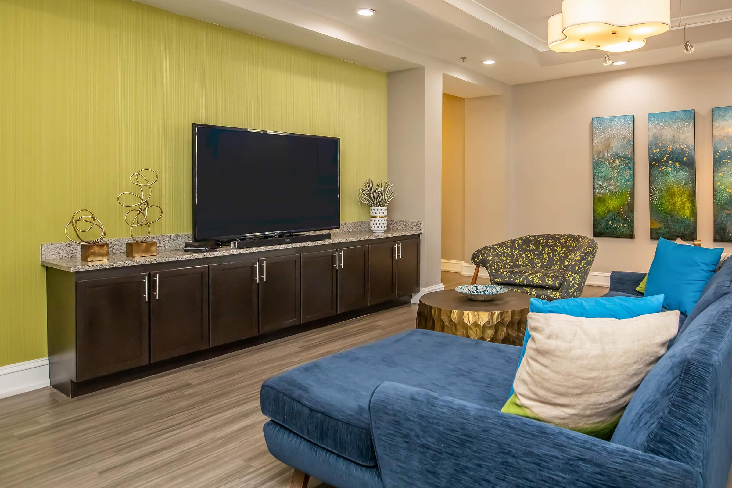 A modern living room featuring a dark blue sectional sofa with colorful pillows, a decorative gold coffee table, and a flat-screen TV mounted on a dark wood cabinet. The wall behind the TV is bright green, and there are three abstract art pieces in blue and green tones hanging nearby.