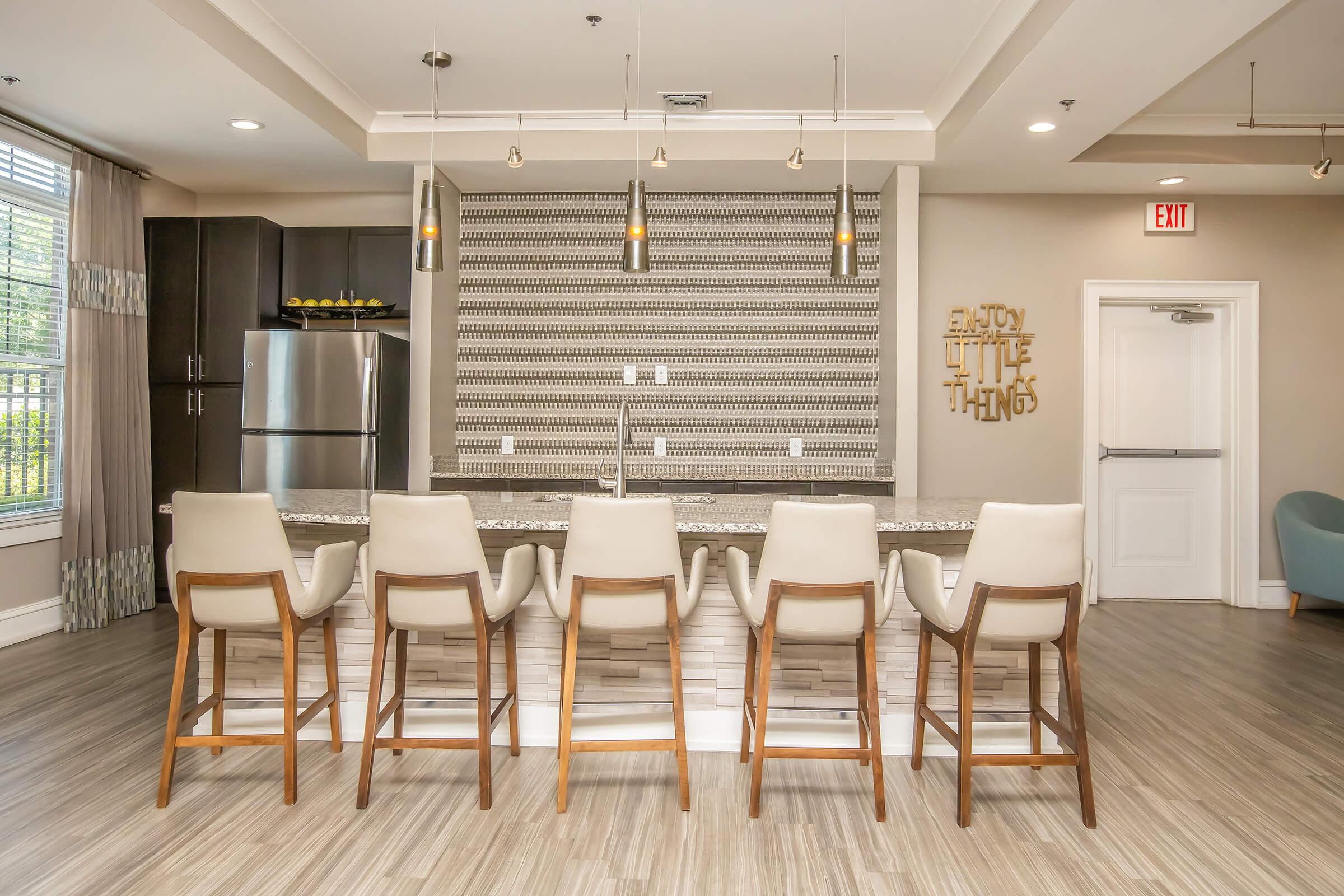 A modern kitchen bar area featuring sleek white and wooden stools arranged in front of a stone counter. The wall behind has decorative tiles, with pendant lights hanging overhead. A stainless steel refrigerator is visible, and a sign that says "Enjoy the little things" is mounted on the wall.